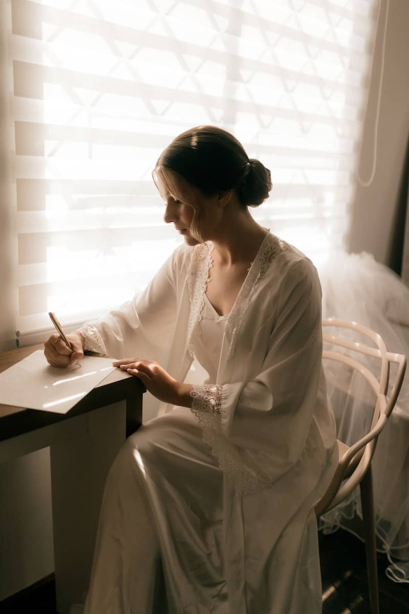 Woman in white dress writing at desk by window