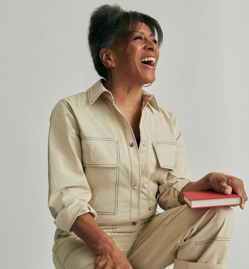 A laughing woman in a jumpsuit holds a red book.