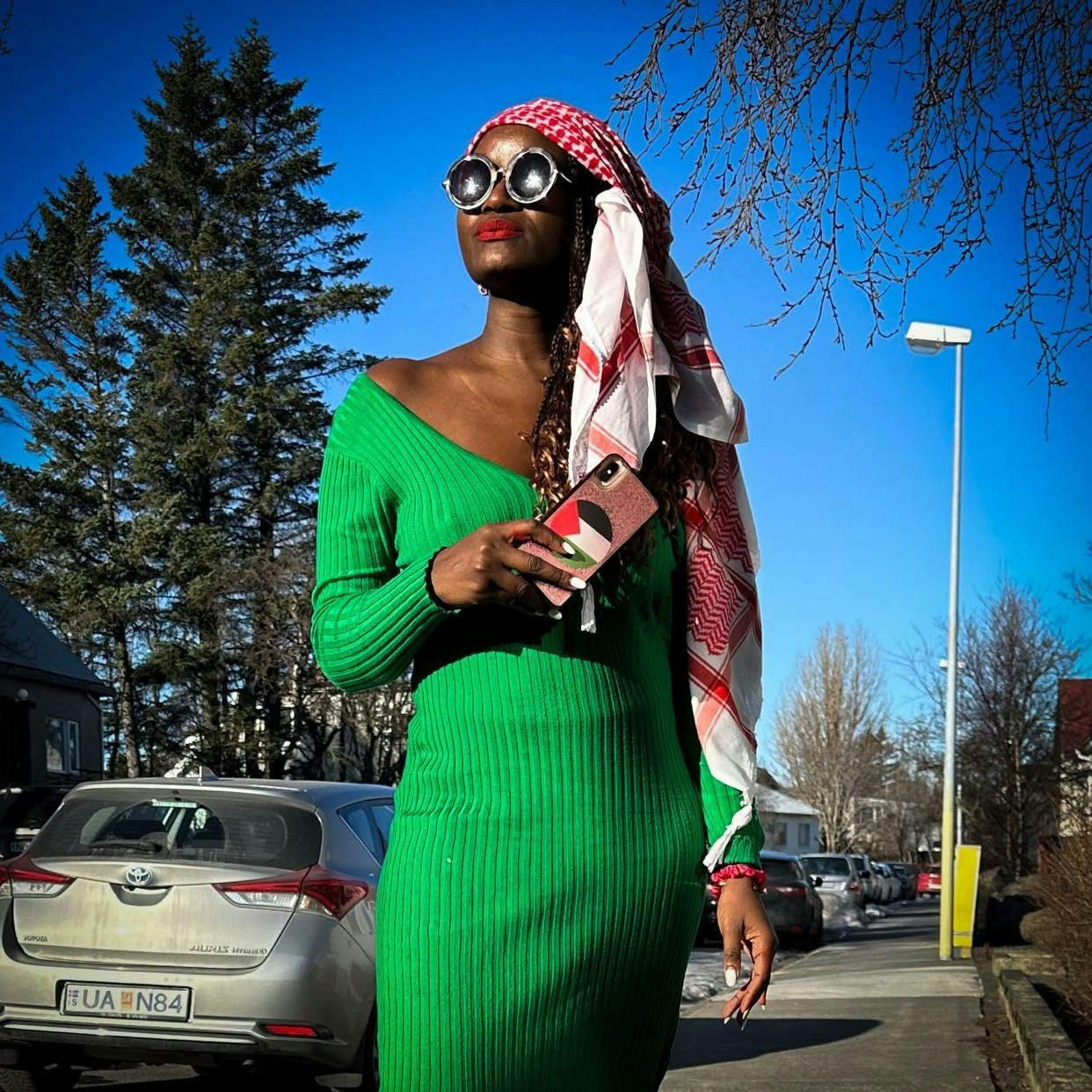  A Black woman, wearing a green dress and sunglasses and a red and white Kaffiye as a headwrap. Holds a phone with a Palestinian flag.