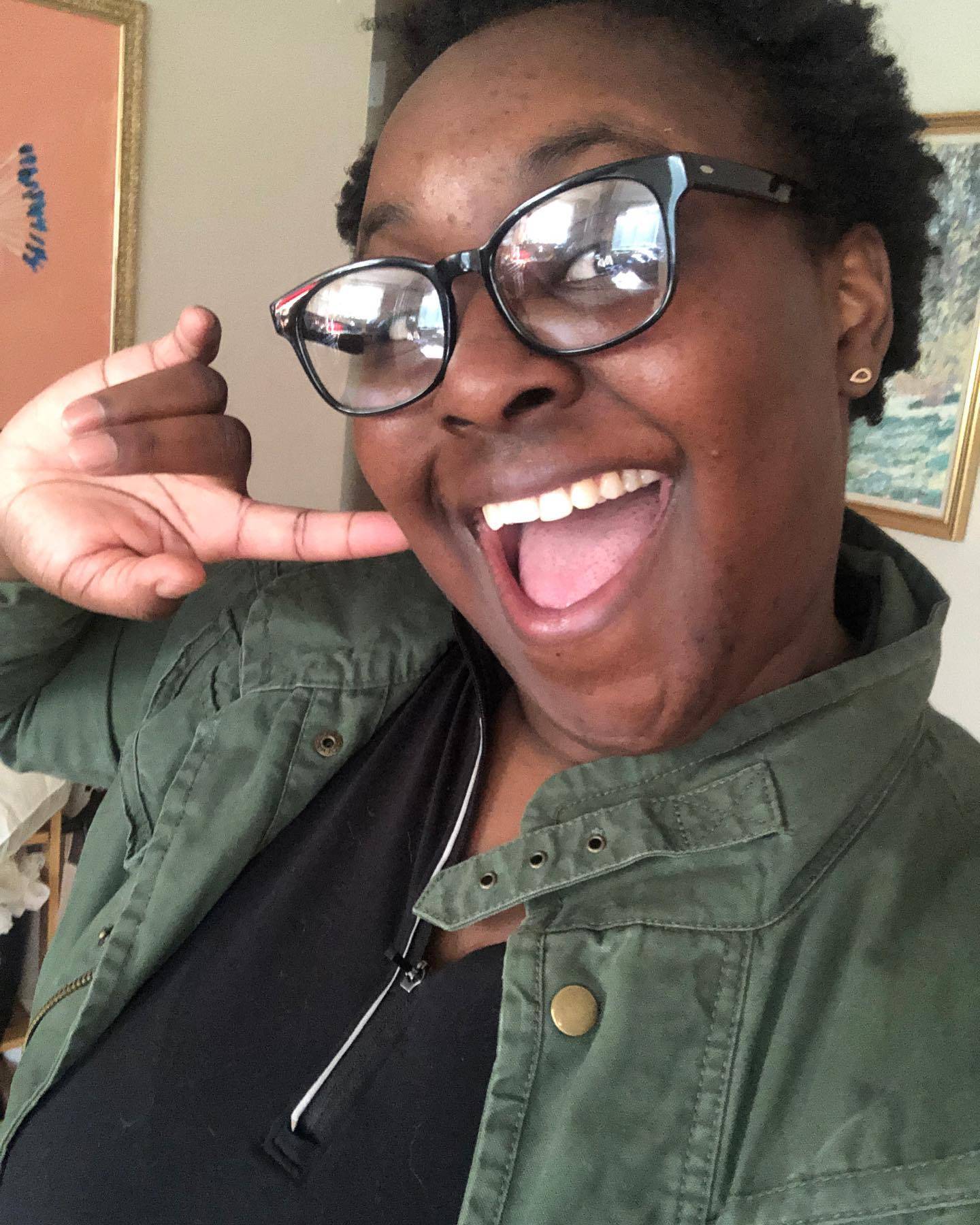 Dark skinned woman with black glasses and a wide open smile. She is wearing a black shirt and green army jacket.