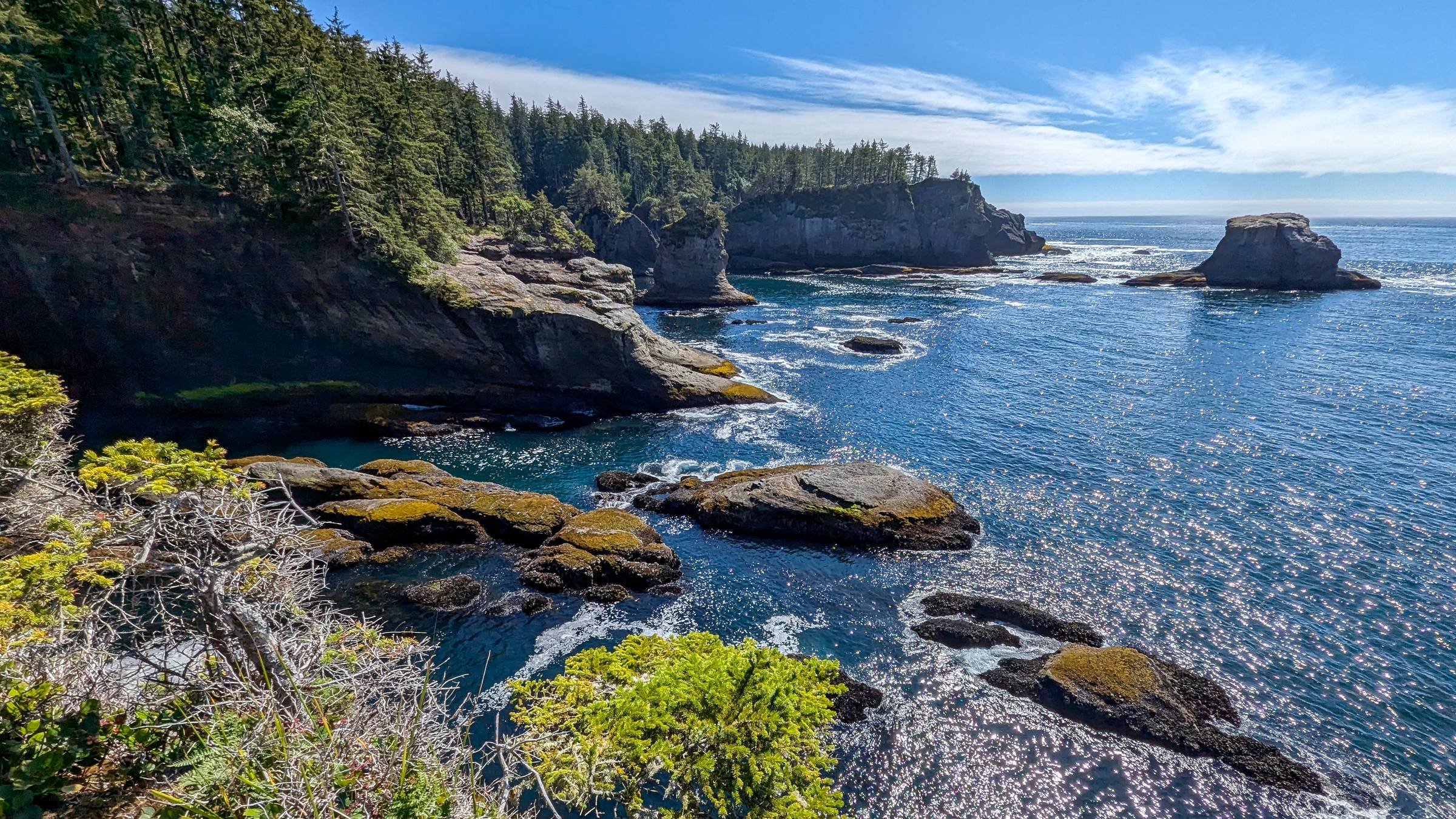 A vibrant coastal landscape at Cape Flattery, showcasing rocky cliffs, lush greenery, and sparkling blue waters. The tide pools and sea stacks are surrounded by shimmering sunlight reflecting off the ocean, creating a picturesque seascape.