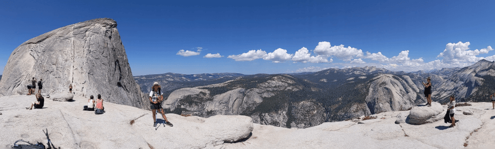 Panoramic view from the summit of Half Dome in Yosemite National Park, showing hikers standing on smooth granite rock surfaces with a steep rock face on the left. In the background, rolling mountain ridges stretch out under a blue sky dotted with clouds, 