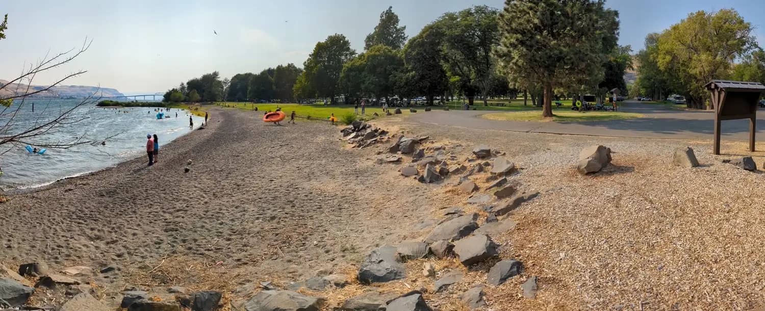 A pebbly beach at Maryhill State Park with people swimming and relaxing near the water, leading into a grassy park area with trees, paths, and picnic shelters.