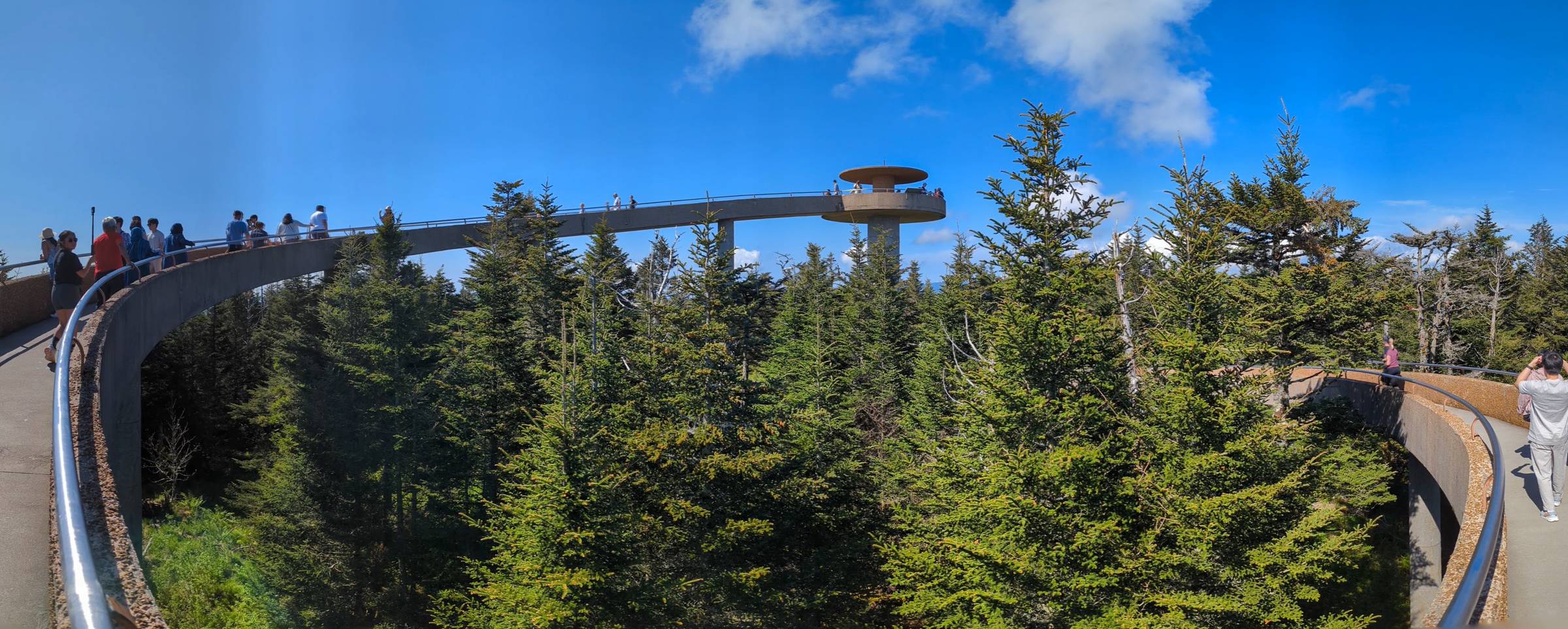 Wide panoramic view of the spiraling concrete ramp leading to Kuwohi Observation Tower, surrounded by a dense evergreen forest under a bright blue sky with scattered clouds. Several visitors walk along the elevated path, enjoying the scenic overlook above