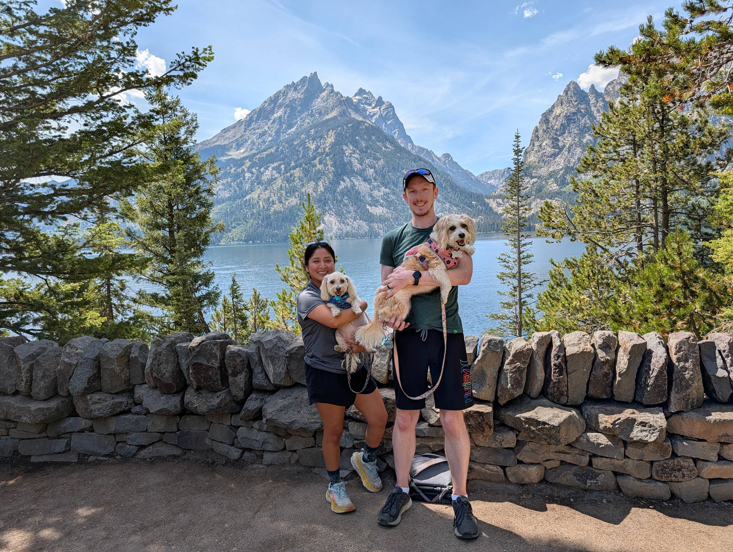 A couple and their two small dogs posing in front of Jenny Lake and the Teton Range at Grand Teton National Park on a sunny day.