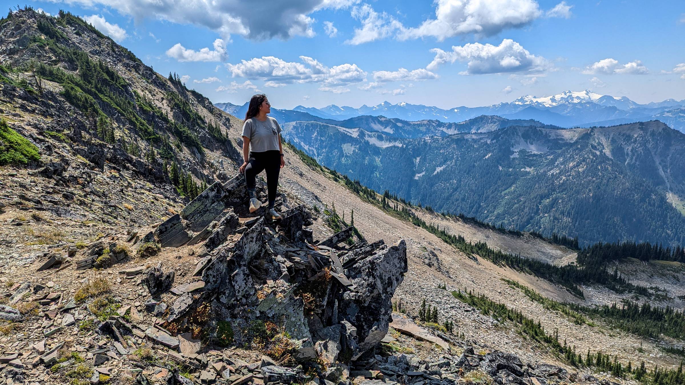 A hiker stands atop a jagged rock outcrop along the Grand Valley Trail, gazing out over a dramatic landscape of forested ridgelines and distant snow-capped peaks, including Mount Olympus under a partly cloudy sky.