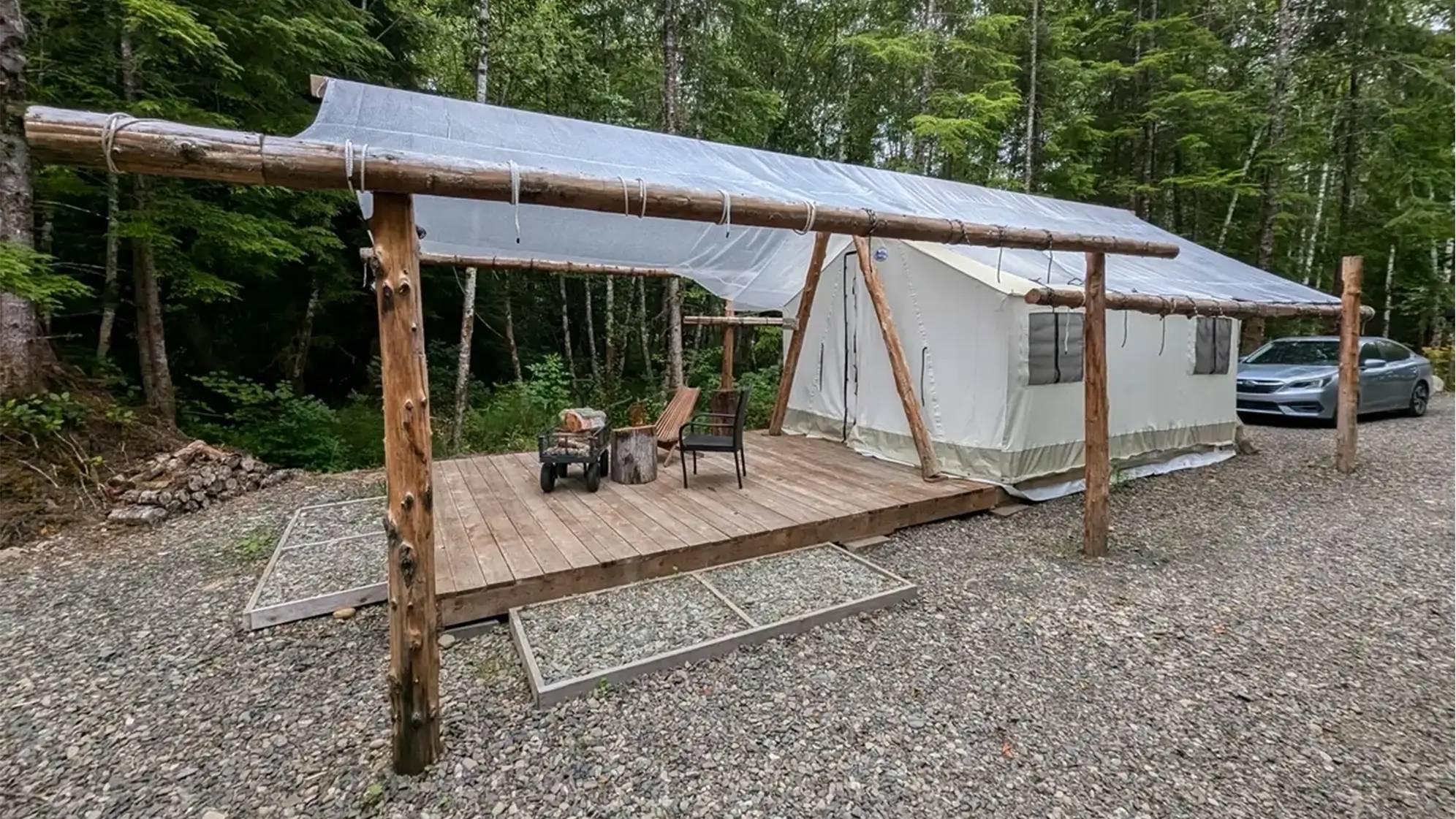 Canvas glamping tent at Mossquatch Resort in Washington’s Olympic Peninsula, set on a wooden deck with a clear rain cover supported by rustic log beams. Surrounded by lush forest, the setup includes a couple of wooden chairs and a small firewood cart, blending comfort with nature.