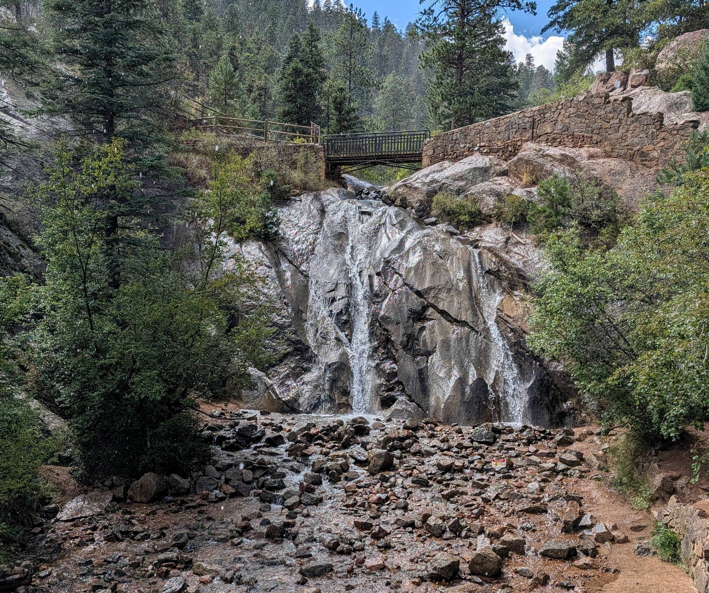Scenic view of Helen Hunt Falls cascading over rocks with a pedestrian footbridge and pine trees under a partly cloudy sky in Colorado Springs.