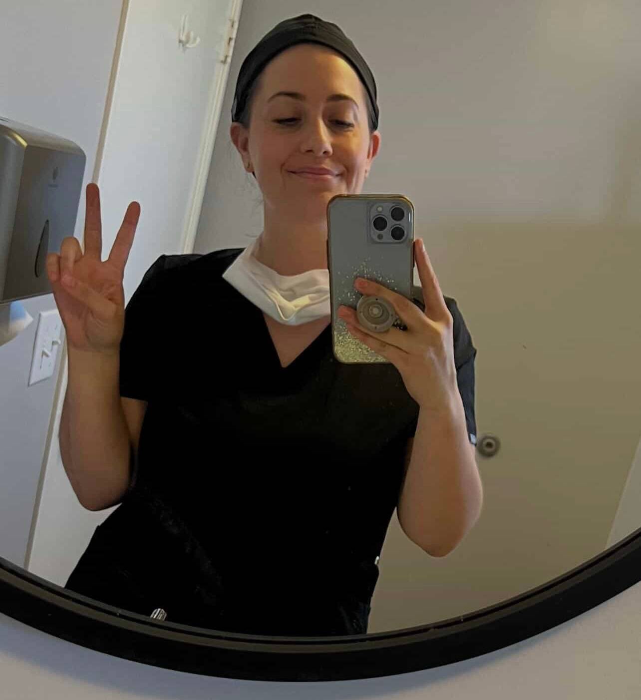 Nurse Shara Hanna is seen in scrubs smiling and making a peace sign with her fingers.