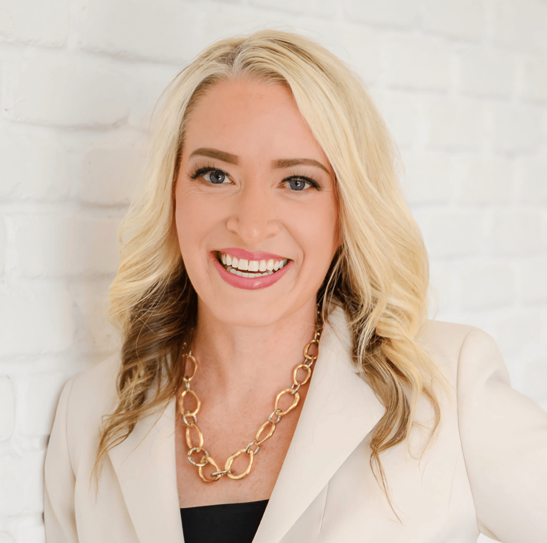 Heather Colledge, a former ICU nurse and current business coach, smiles while wearing gold jewelry and a white blazer.
