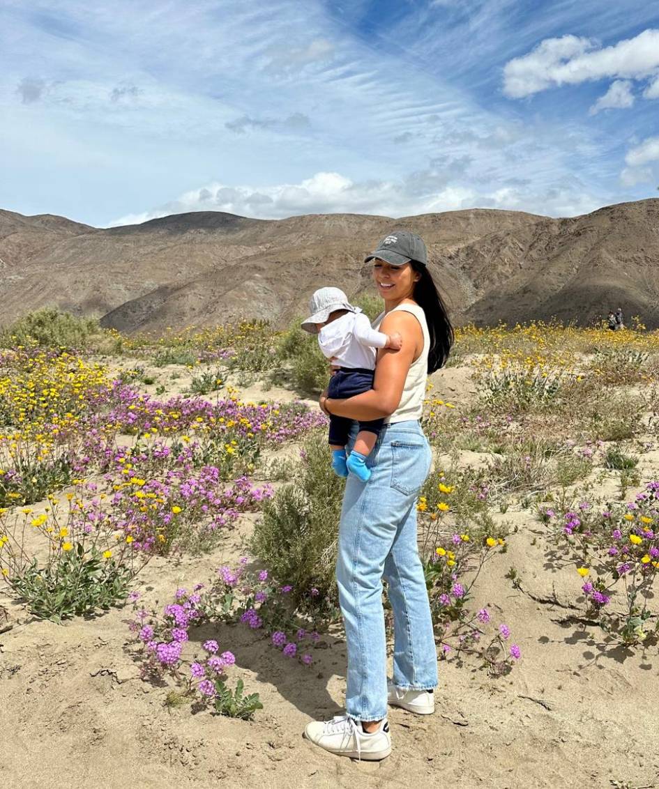 Woman standing in a wildflower field holding a baby, with mountains in the background.