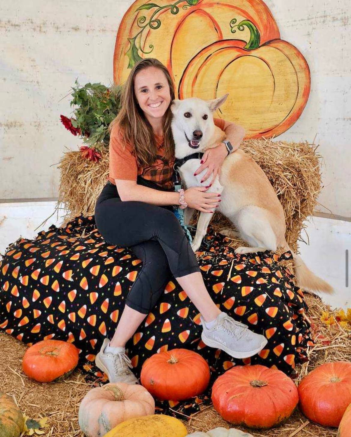 A nurse sitting on a fall-themed blanket with her dog, surrounded by pumpkins and hay bales.