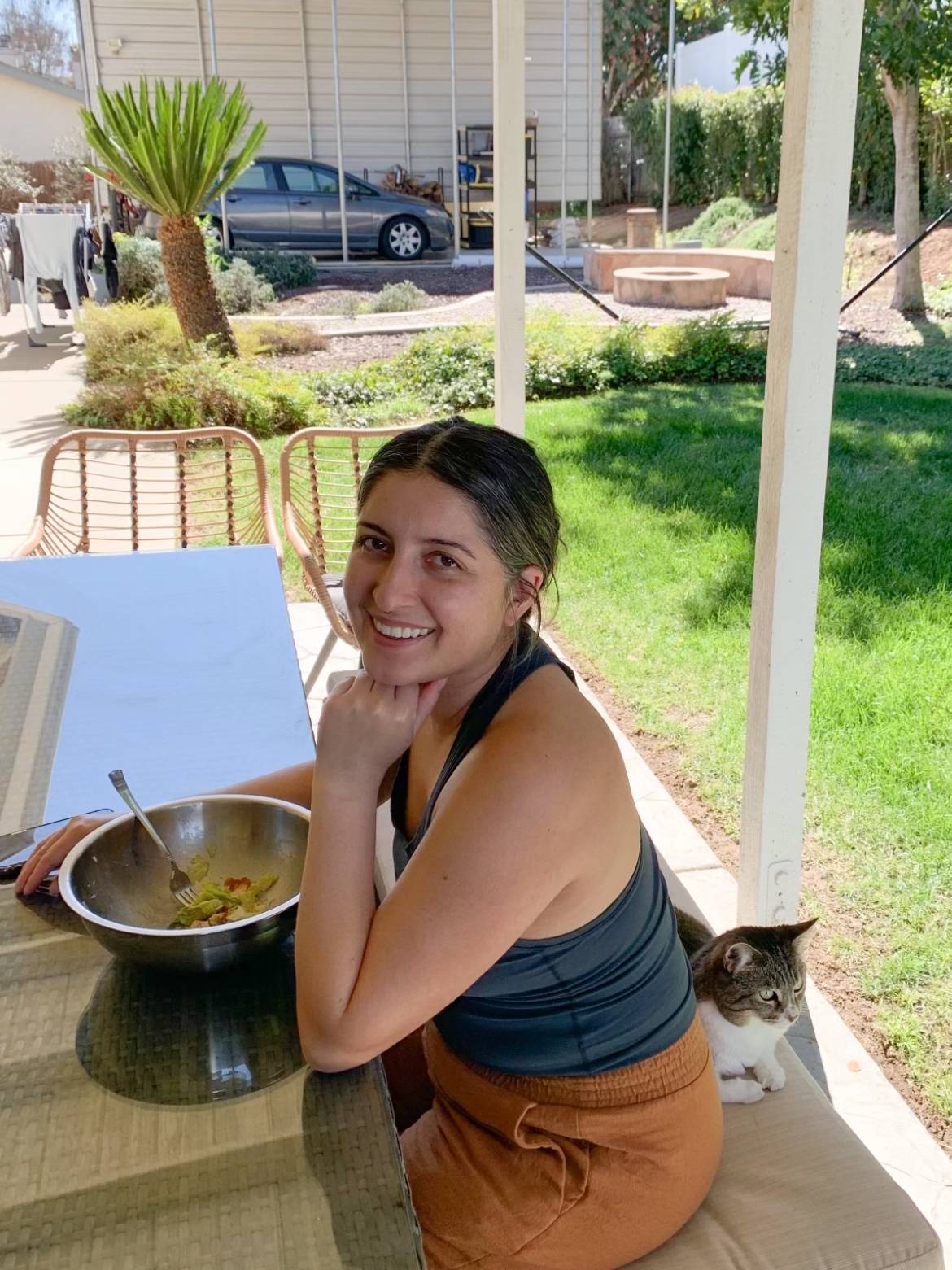 Jackie Contreras, RN, BSN, RNC-NIC, CLC, a neonatal intensive care nurse, smiles in a backyard while eating salad and sitting next to a cat.