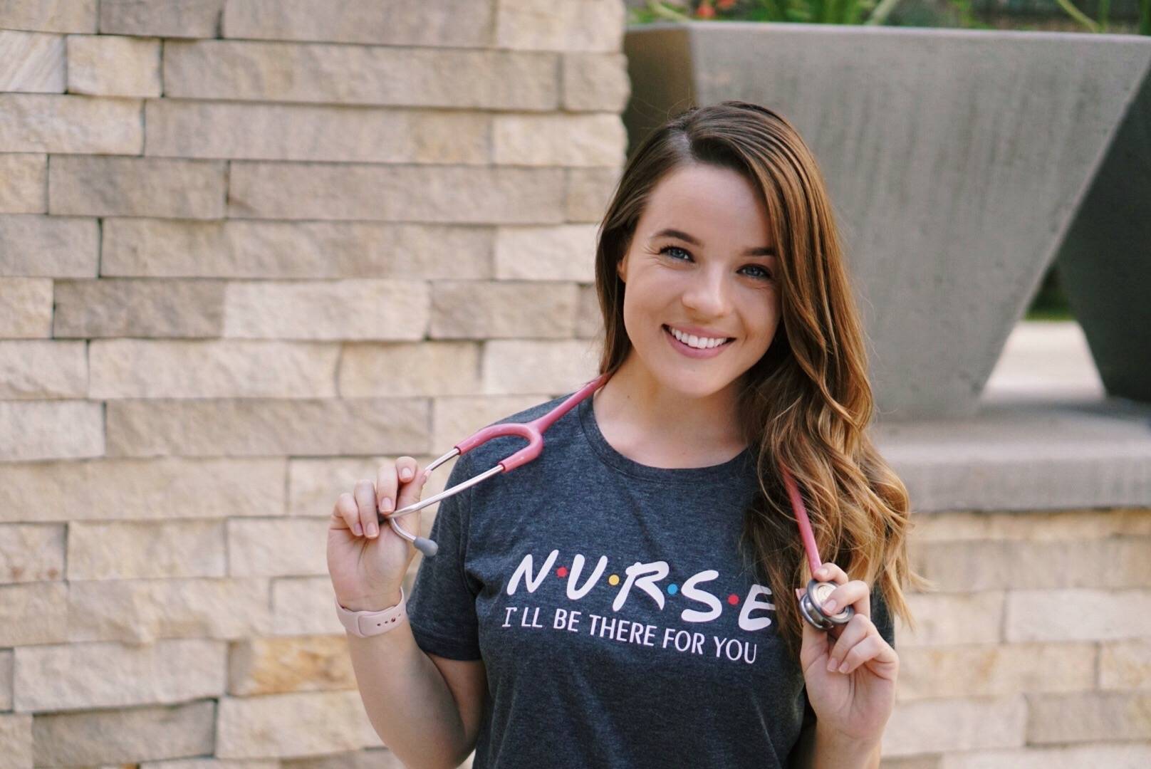 A smiling nurse holding a backpack strap while wearing a “Nurse” shirt outdoors.