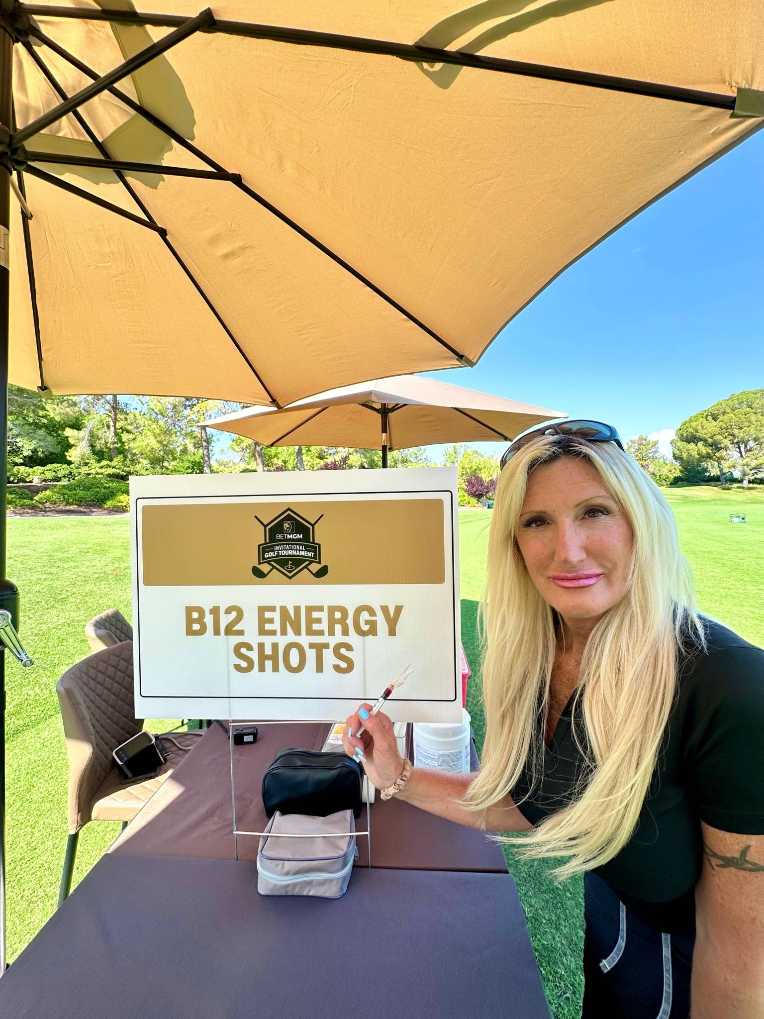 Person sitting outdoors under an umbrella holding a sign that says “B12 Energy Shots.”