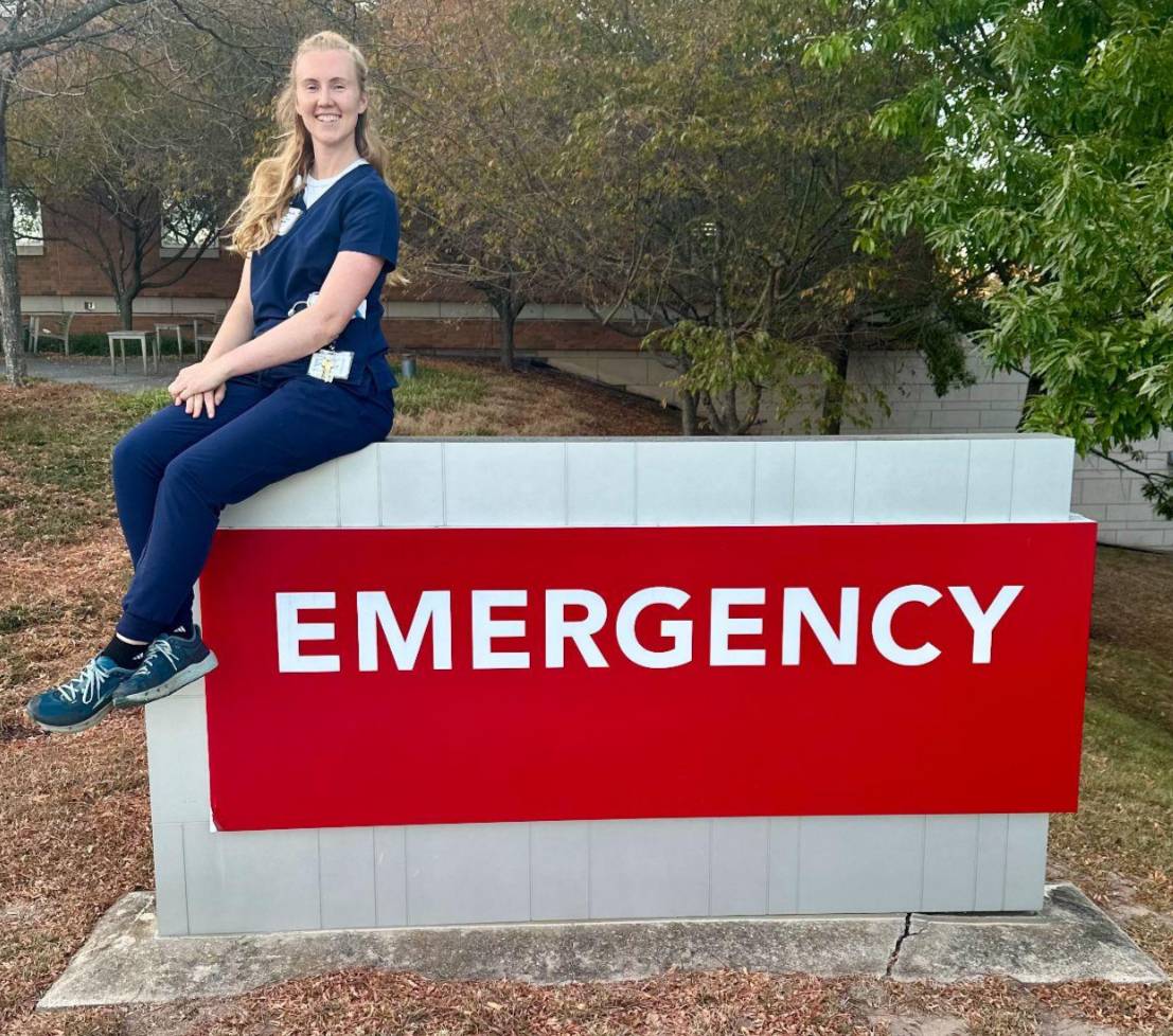 Nurse in scrubs sitting on an emergency sign.