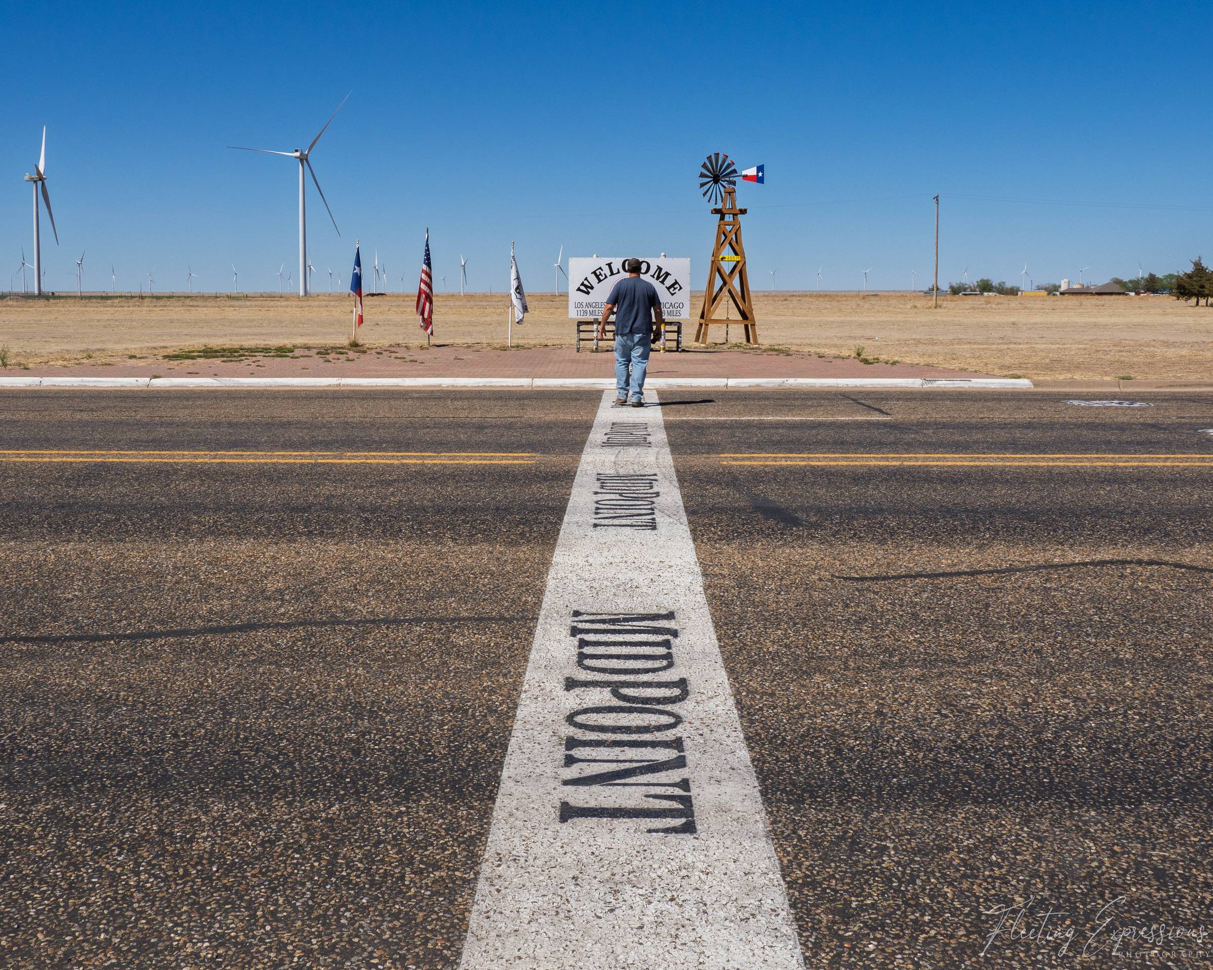 A man walks down the painted center stripe of Route 66 toward a midpoint welcome sign in Adrian, Texas, with wind turbines visible on the flat horizon.