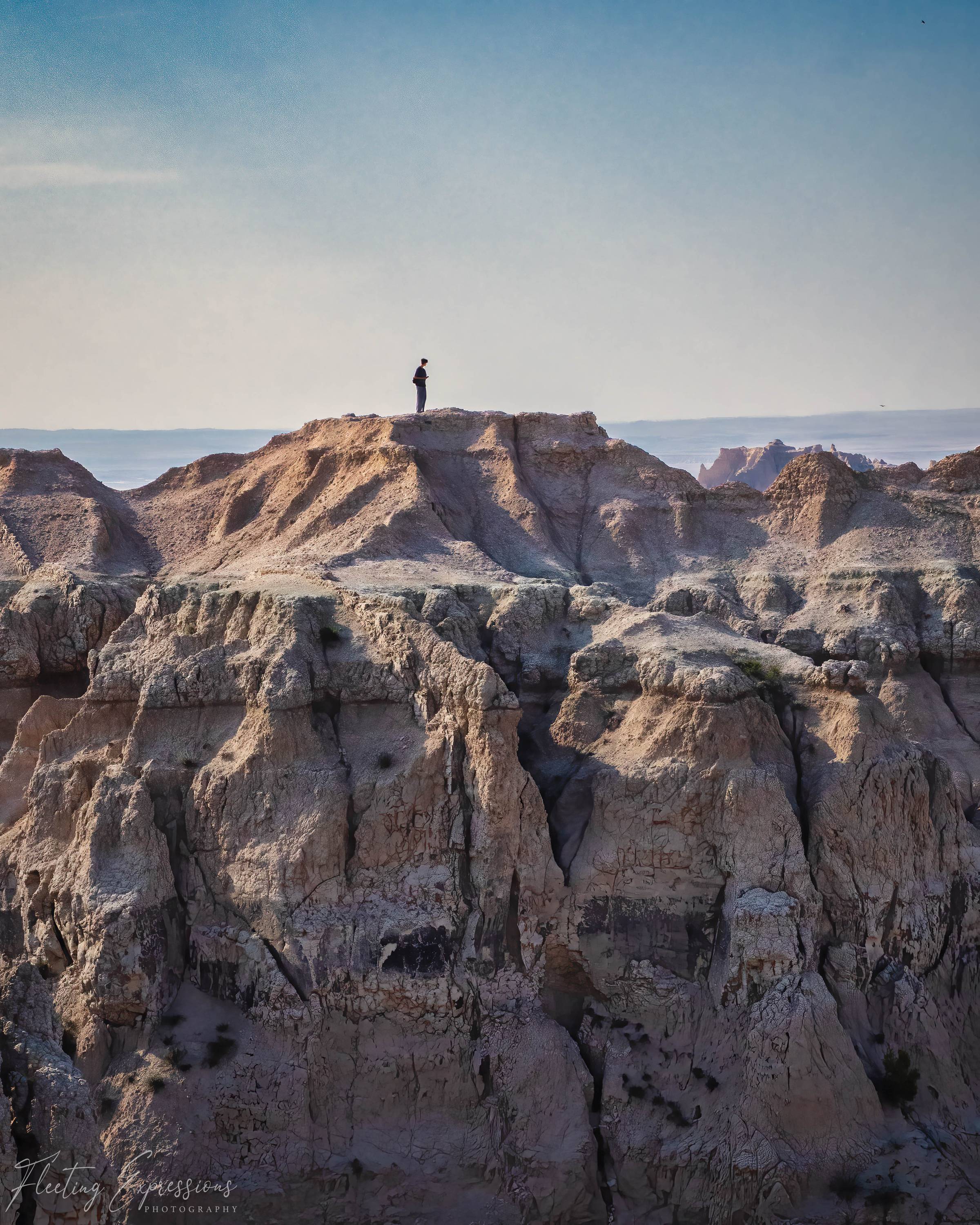 Small figure standing at the edge of a rugged badlands rock formation, silhouetted against a pale blue sky with layered rock formations stretching into the distance.