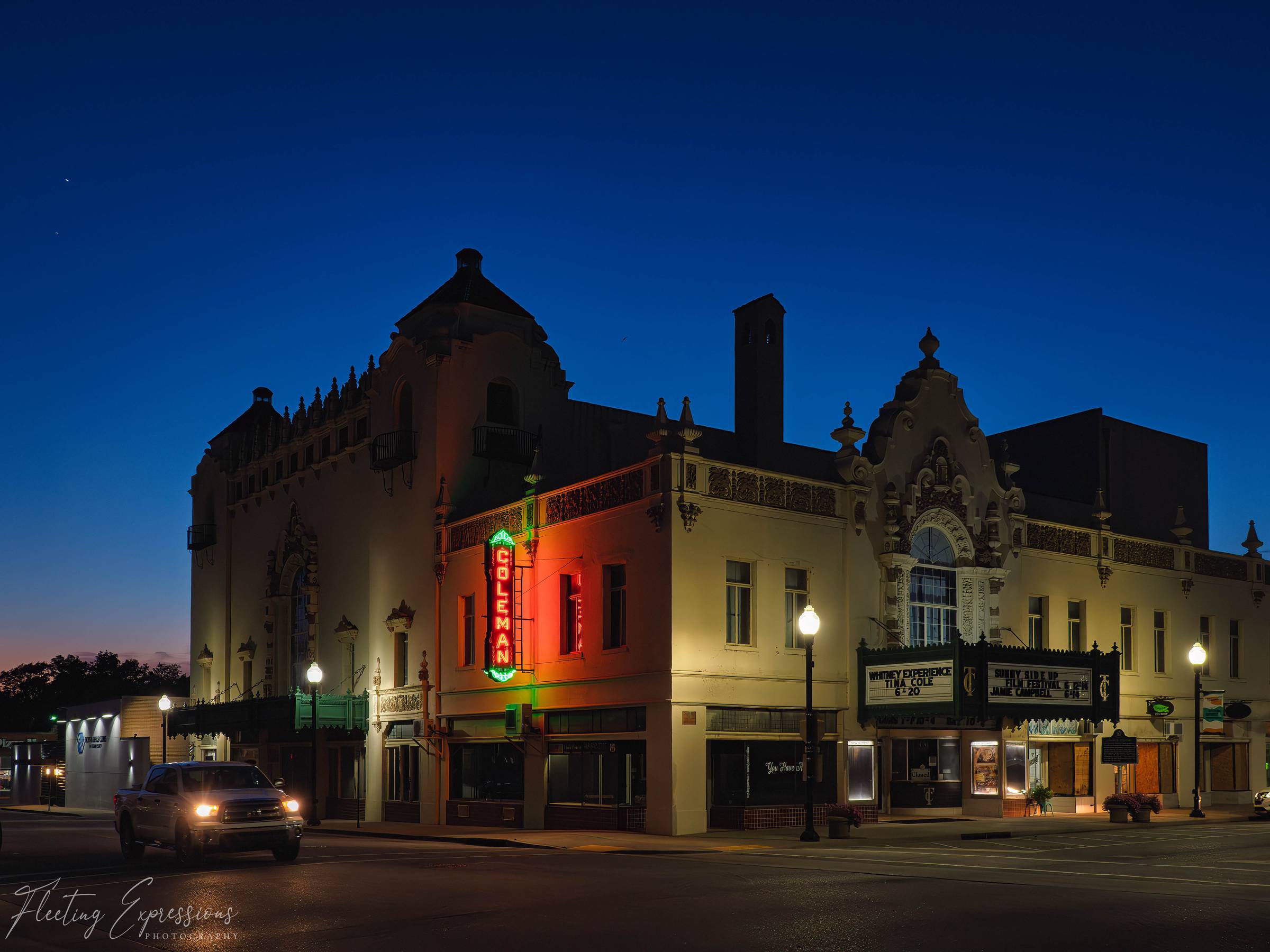 The historic Coleman Theatre glows with colorful neon lights at dusk in downtown Miami, Oklahoma, beneath a deep blue evening sky.