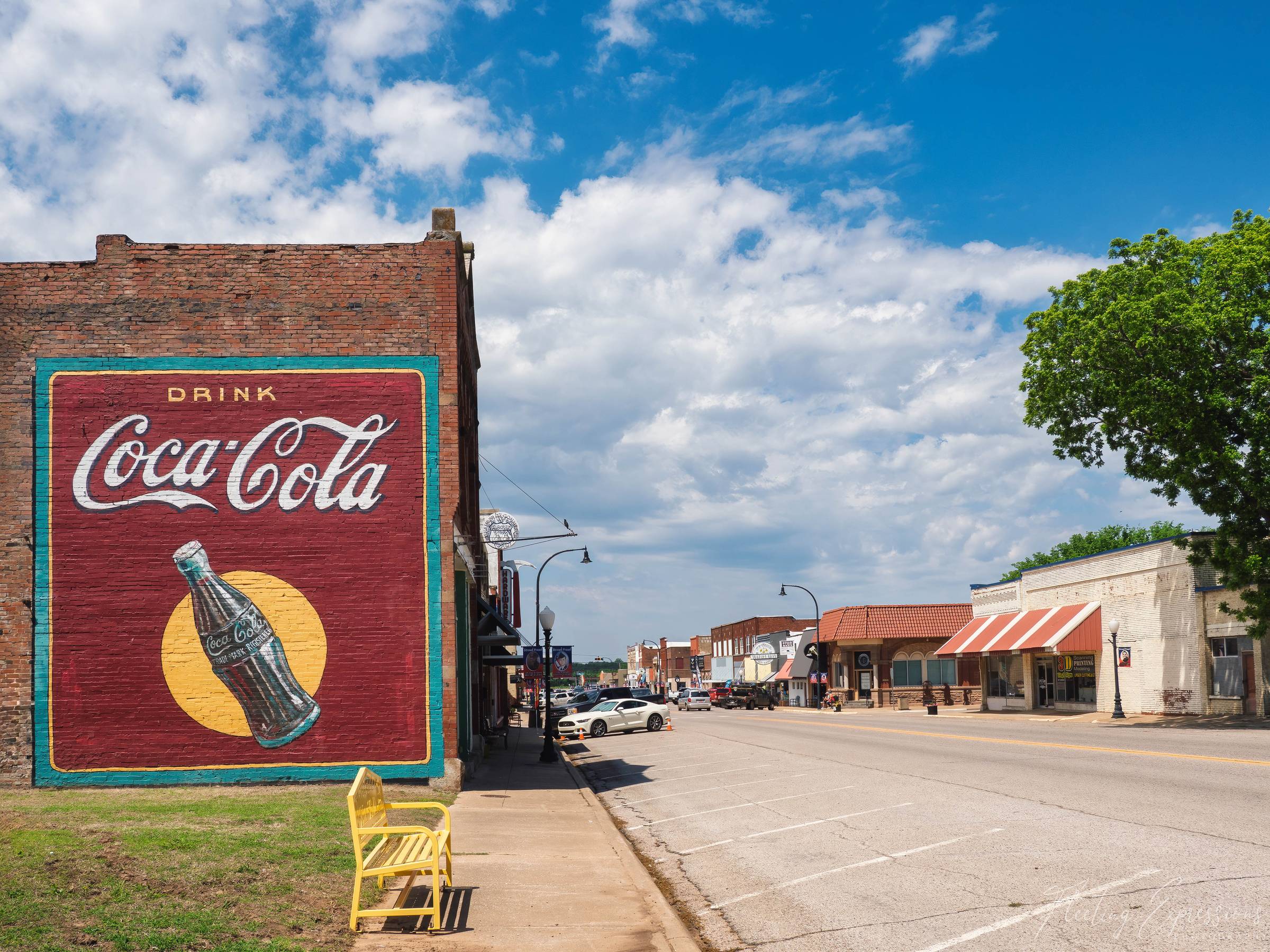 A large vintage Coca-Cola mural painted on the brick side of a building anchors the corner of downtown Stroud, Oklahoma, with the historic main street stretching into the distance under a blue sky with white clouds.