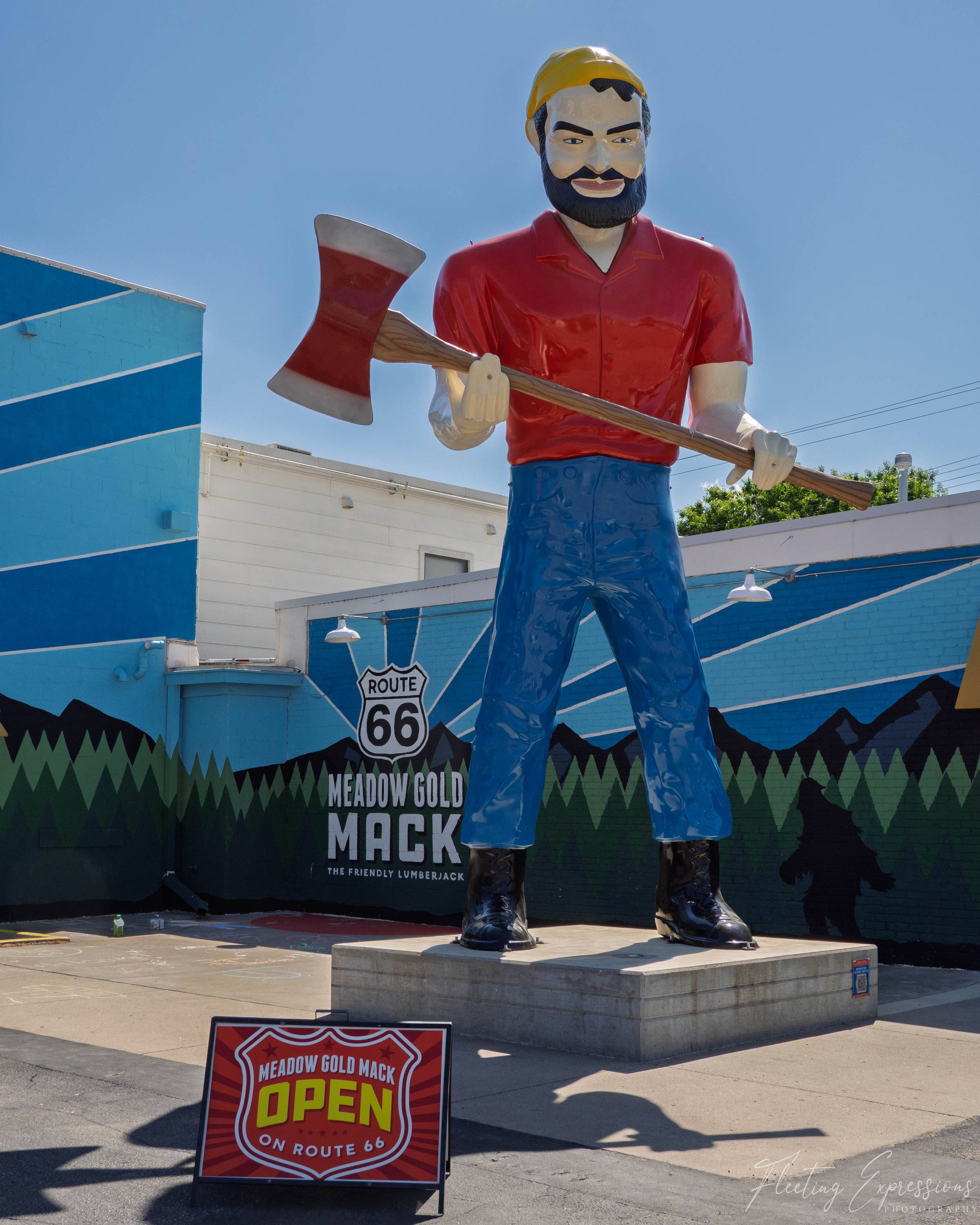 A giant lumberjack statue in a red shirt holds an axe across his body, standing before a forest mural reading “Meadow Gold Mack.” 