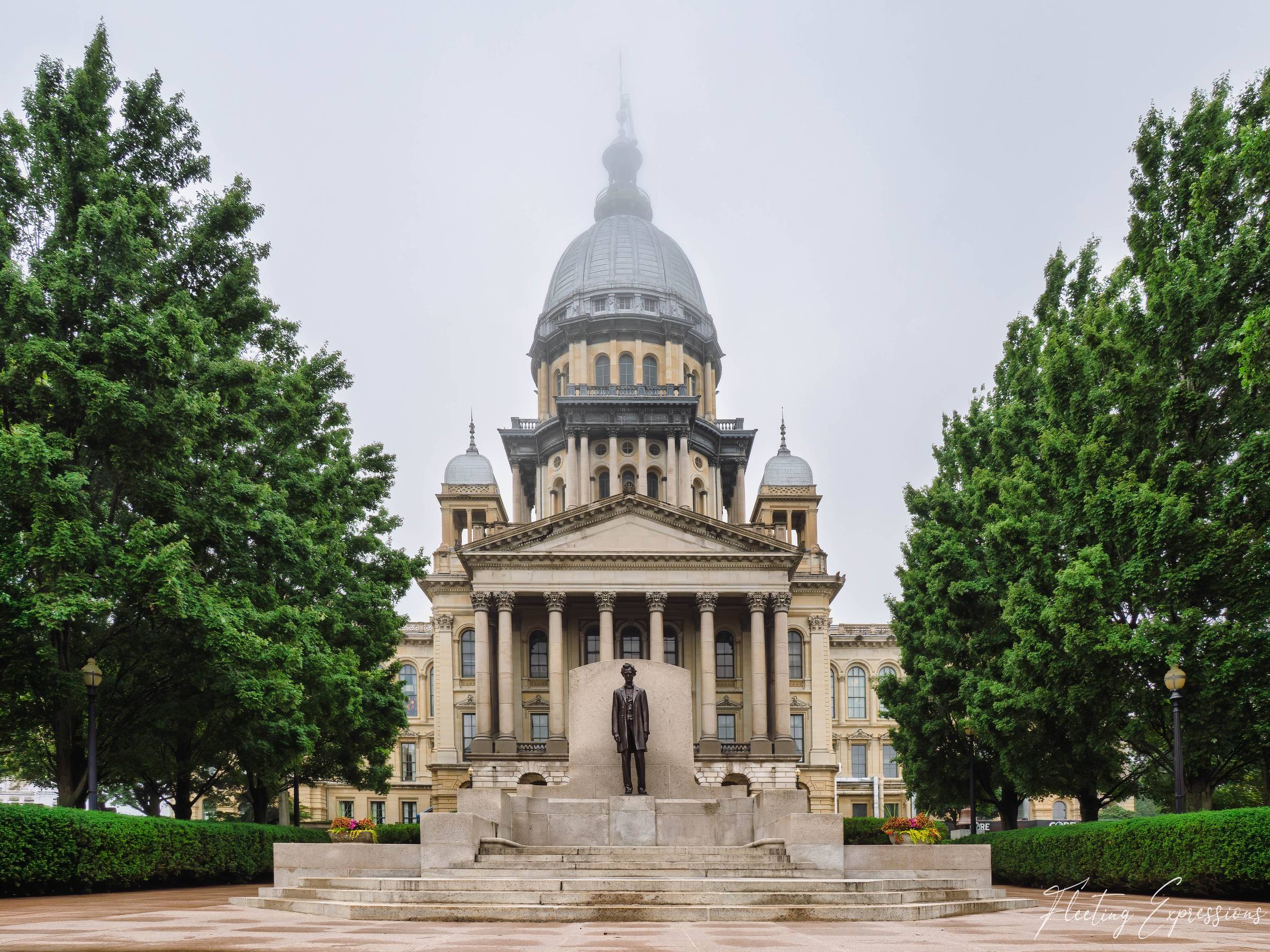 The Illinois State Capitol dome disappears into fog above a bronze Lincoln statue on the plaza; tall green trees frame both sides symmetrically.