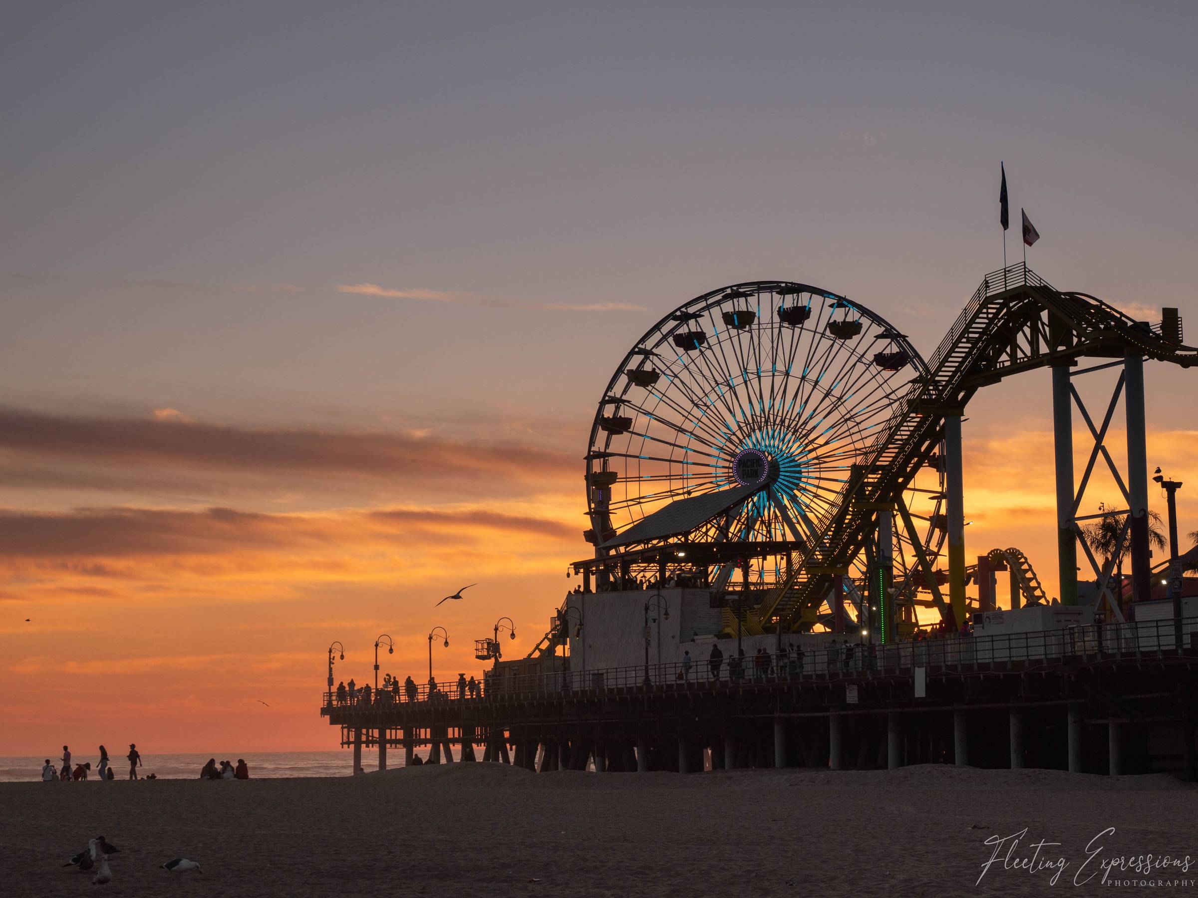 Silhouette of the Santa Monica Pier at sunset, with the Pacific Park ferris wheel and roller coaster against a deep orange sky and small figures walking on the beach below.