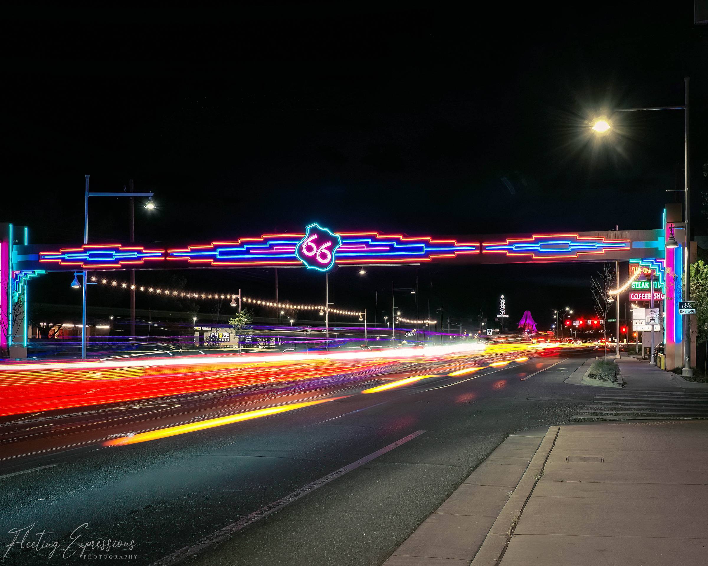 The Route 66 neon arch over Central Avenue in Albuquerque, New Mexico, at night with colorful car light trails streaking beneath it.