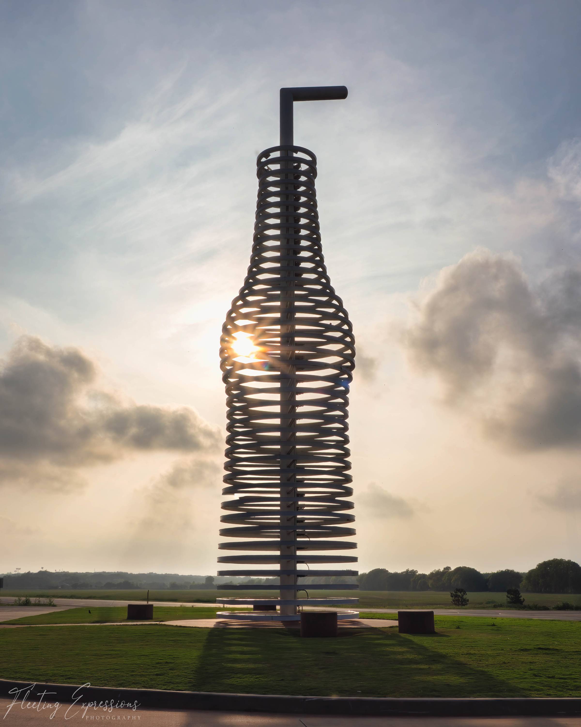 A large steel sculpture shaped like a soda bottle with a straw, constructed from open horizontal rings, backlit by a golden sunburst at sunset in Arcadia, Oklahoma along Route 66.