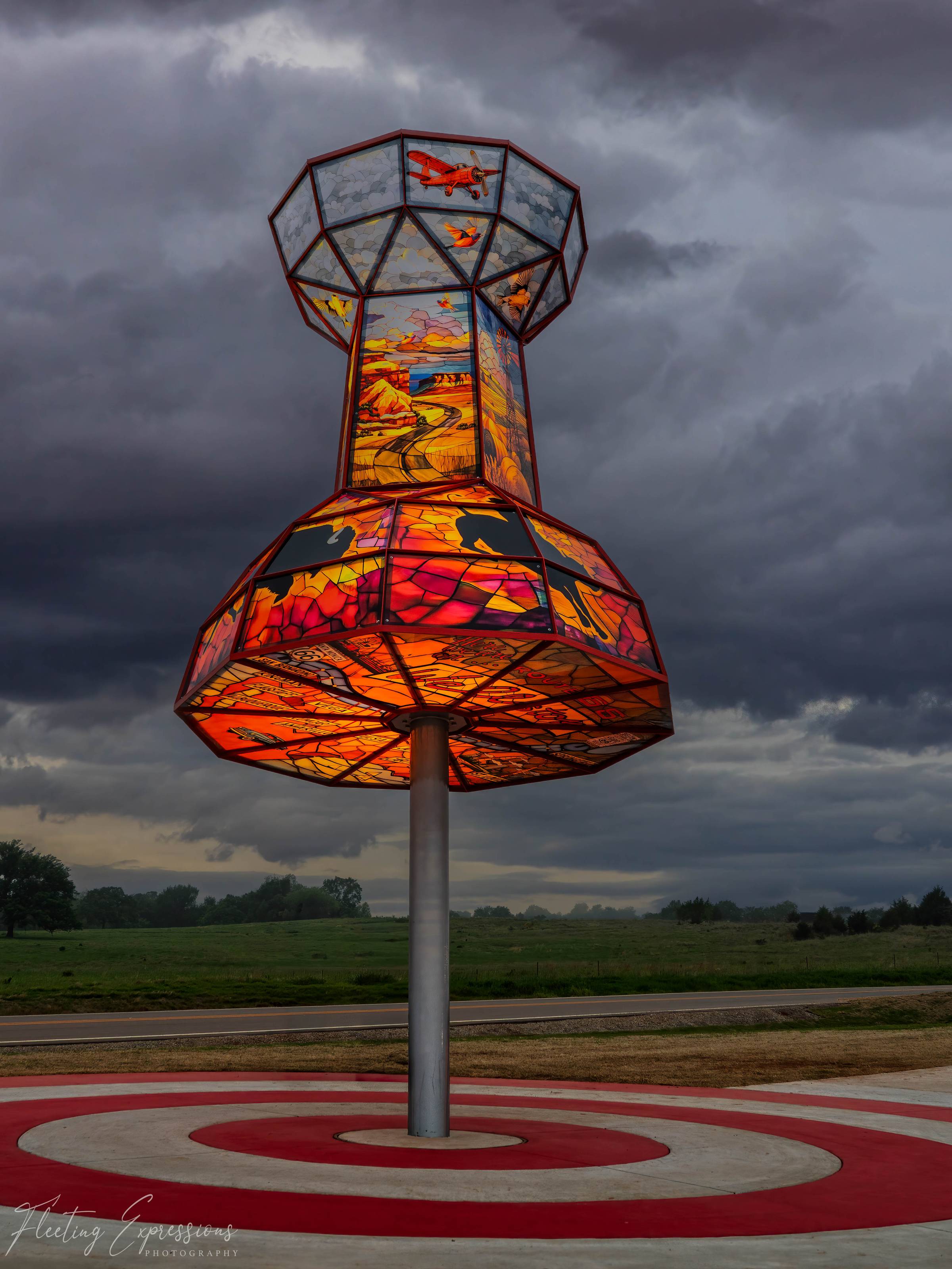 A large illuminated sculpture shaped like a map pin, constructed from colorful stained glass panels depicting Oklahoma history, standing against a dramatic stormy sky in Wellston, Oklahoma.