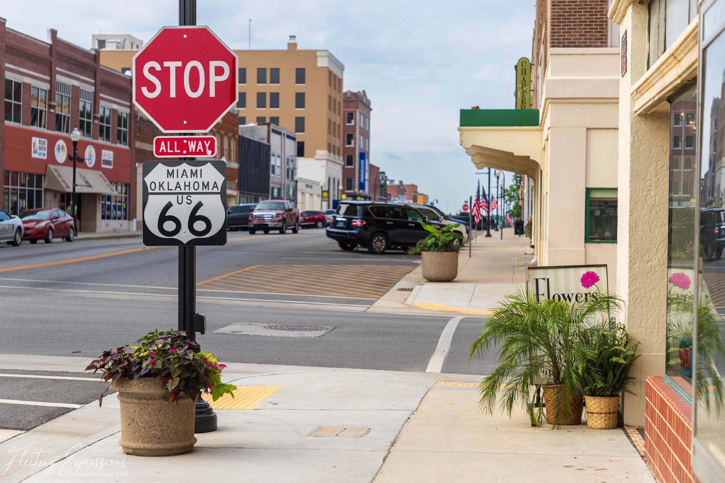 Route 66 highway sign mounted beneath a stop sign along a quiet downtown street in Miami, Oklahoma, with storefronts and parked cars in the background.