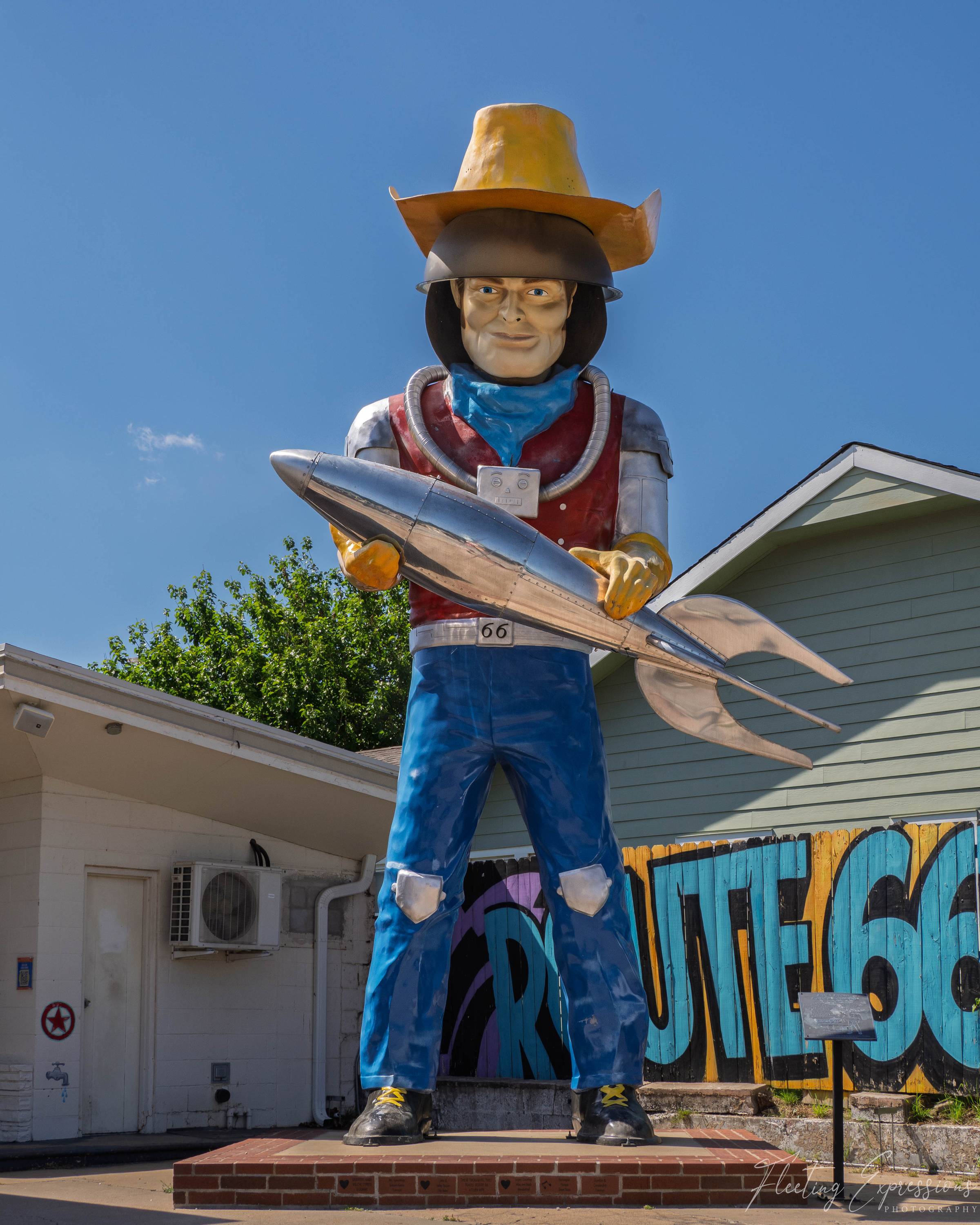 A towering cowboy statue in patched blue jeans and a yellow hat cradles a chrome rocket across both arms beneath a clear blue sky. 