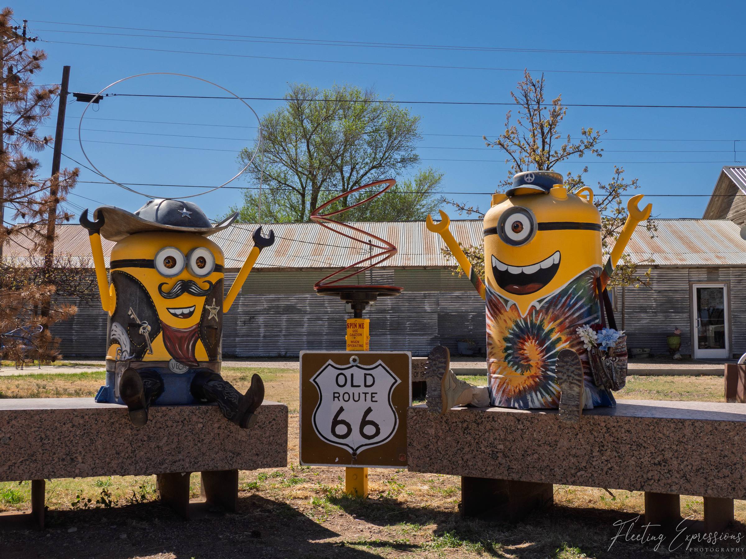 Two painted Minion sculptures dressed as a cowboy and a hippie sit on a bench beside an Old Route 66 sign in Vega, Texas.