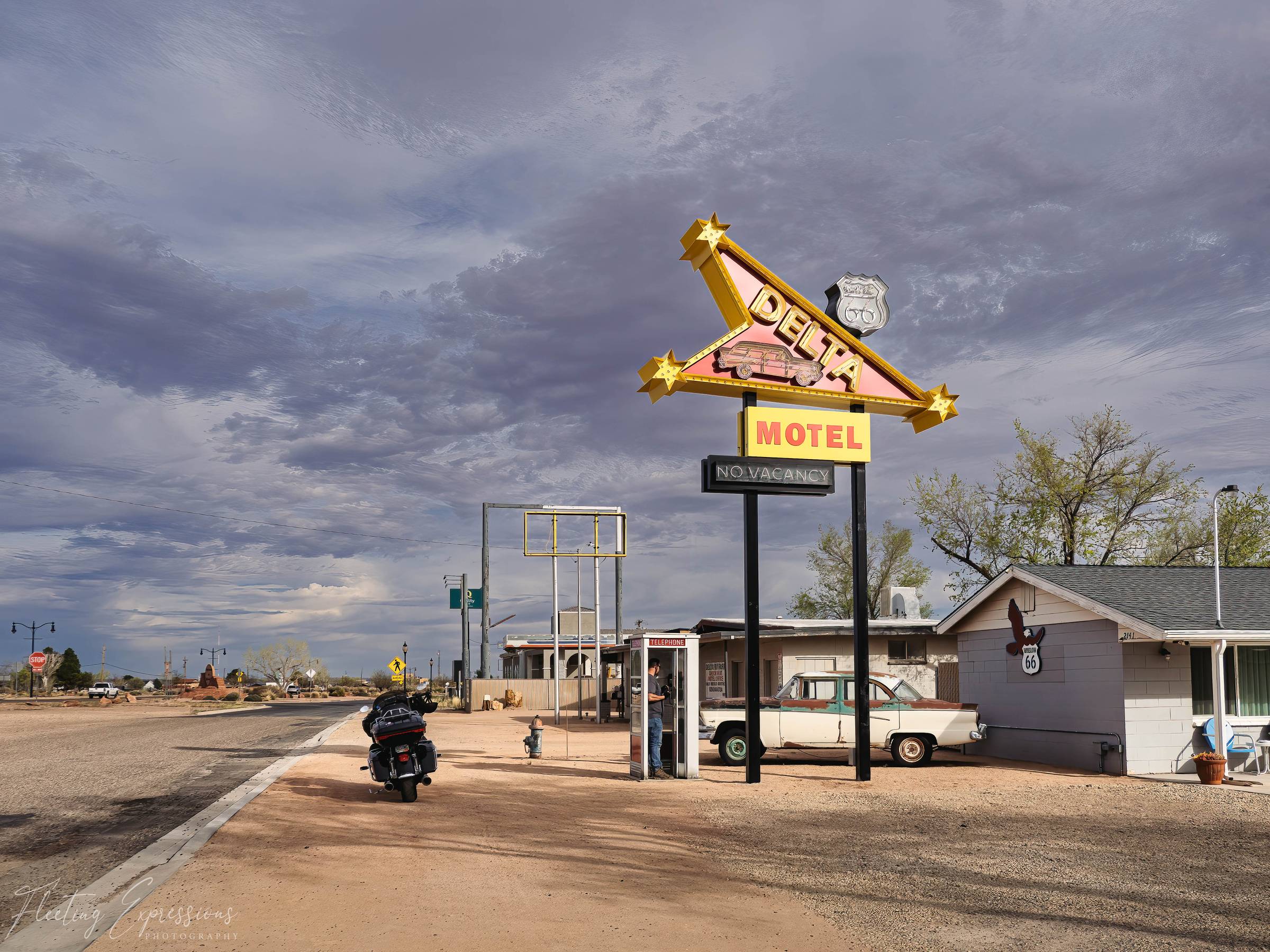 The Delta Motel sign with stars and a Route 66 shield glows against a stormy sky in Winslow, Arizona, with a loaded Harley parked out front and a figure in a phone booth nearby.