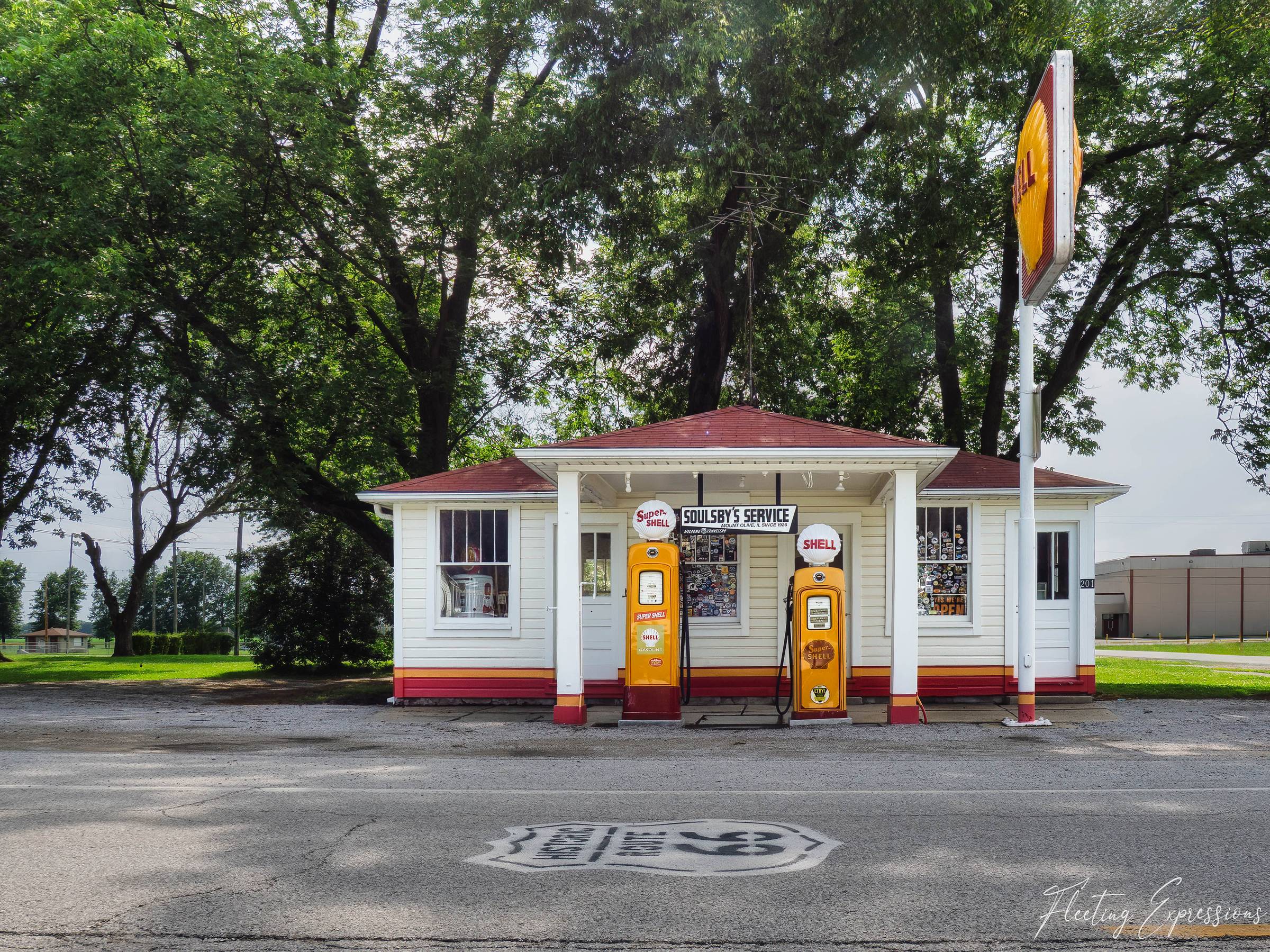 A small white Shell service station with red roof and columned porch sits behind two yellow Super Shell gas pumps; a Route 66 shield is painted on the asphalt in the foreground.