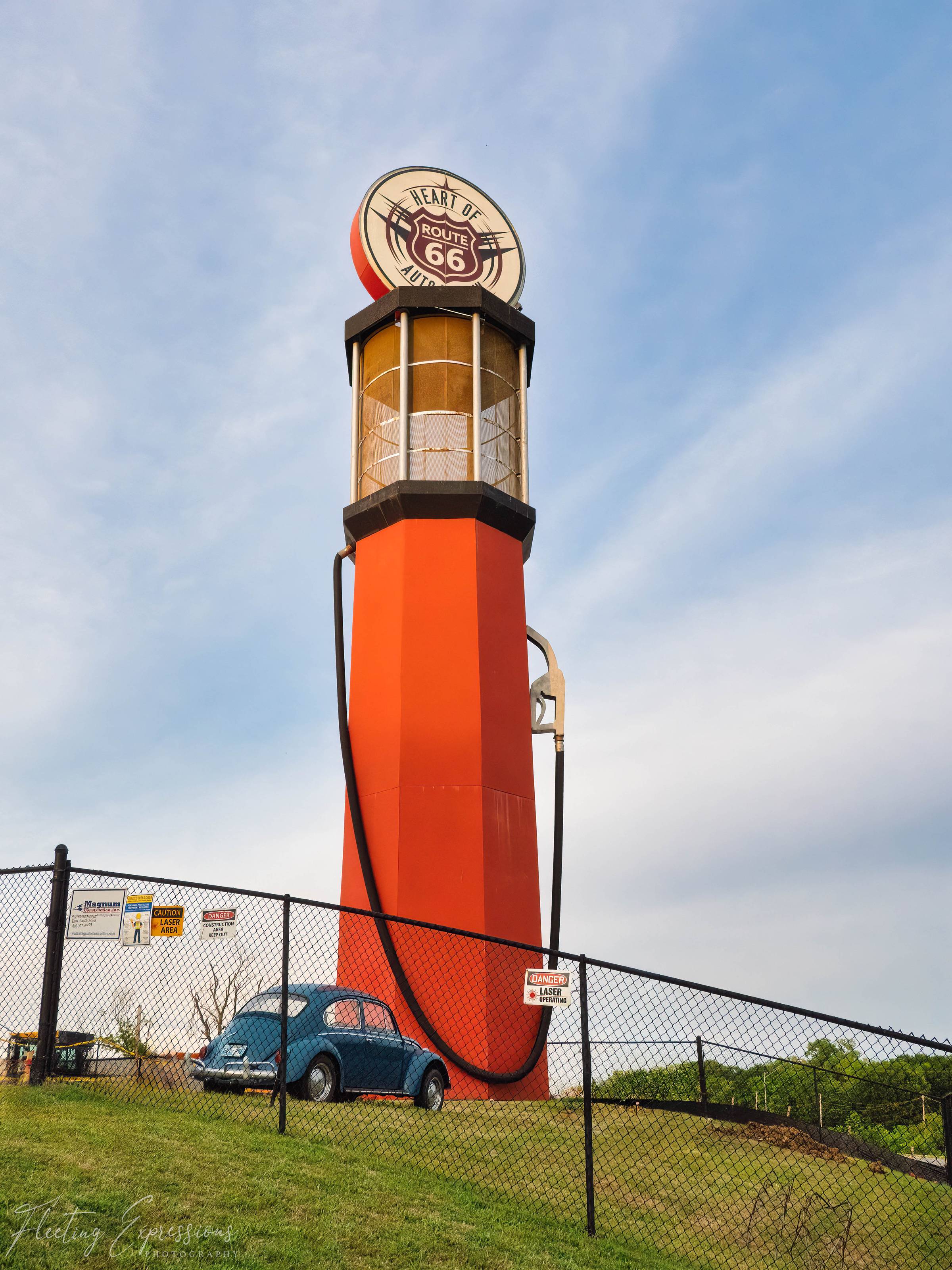 Towering red Route 66 gas pump sculpture behind a chain-link fence, with a small blue VW Beetle parked at its base for scale.