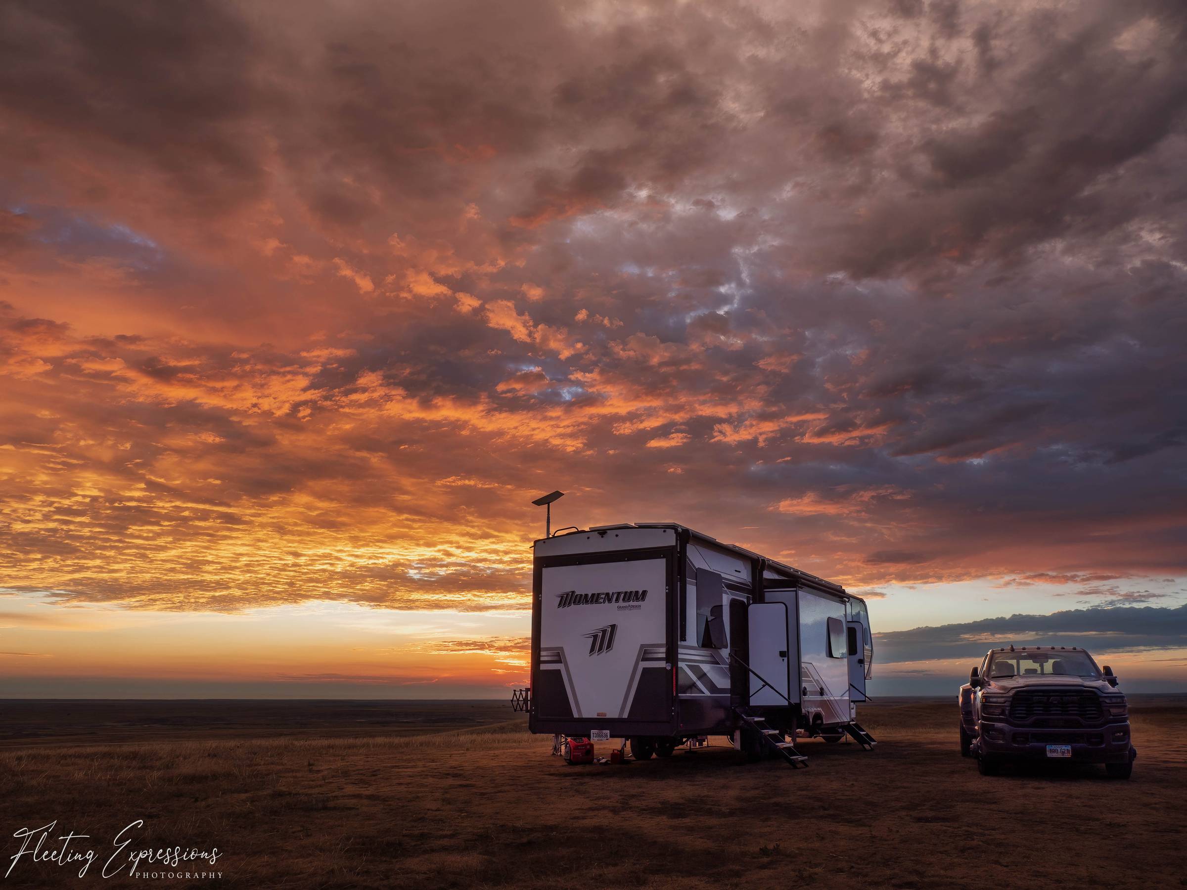  Fifth wheel RV and pickup truck parked in open grassland at sunrise, with orange and pink clouds filling the sky above the Badlands.