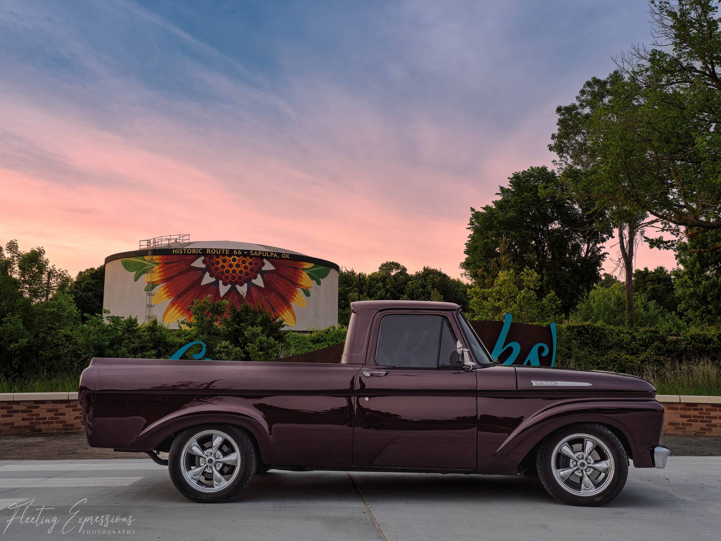 Dark maroon vintage Ford F-100 pickup parked in profile in front of the Get Your Kicks sign and the Indian Blanket water tower at sunset.