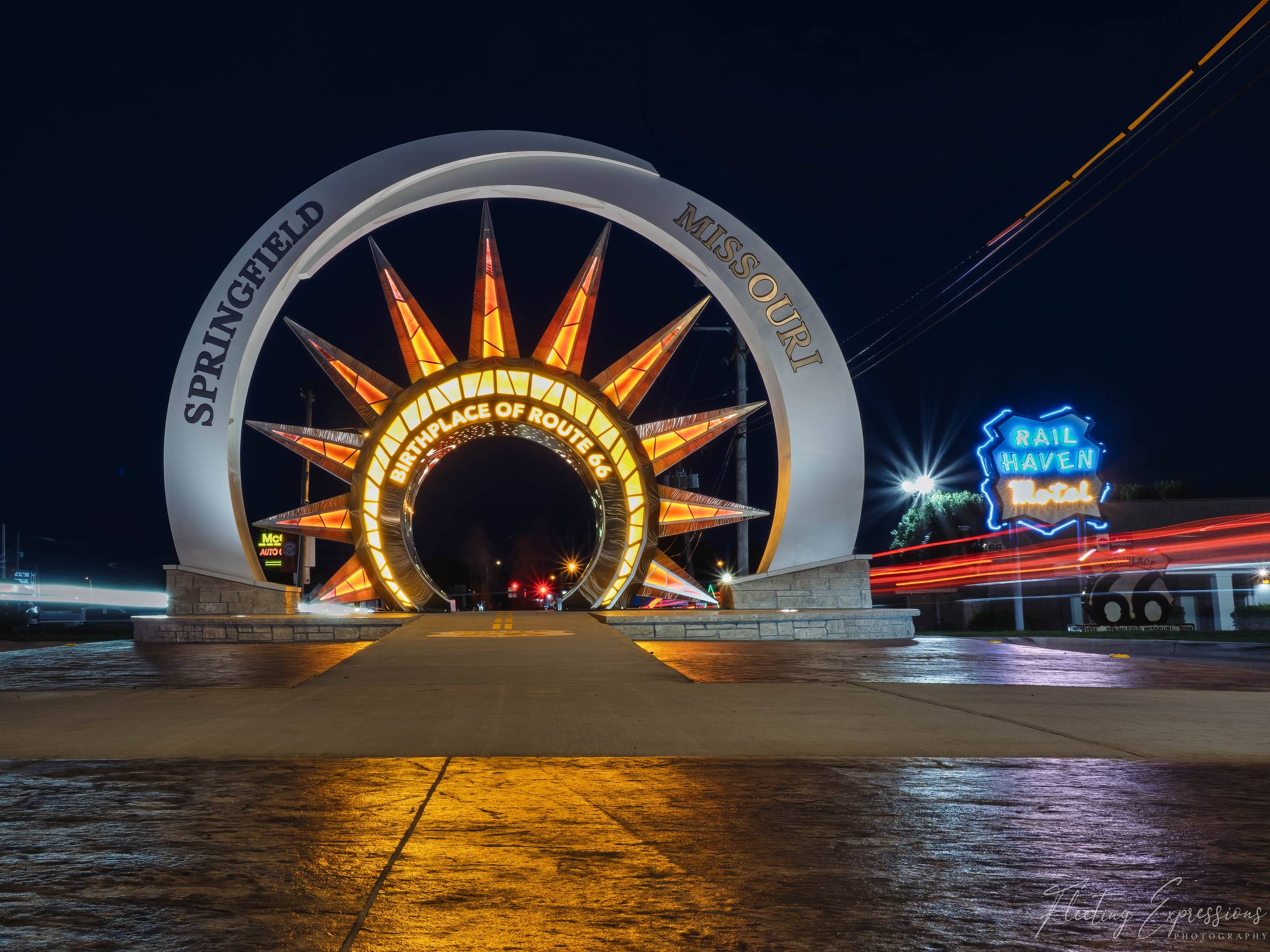 An illuminated 20-foot arch with a sunburst center reading "Birthplace of Route 66" stands on St. Louis Street at night, with the Rail Haven Motel neon sign glowing to the right and red taillight trails across the pavement.