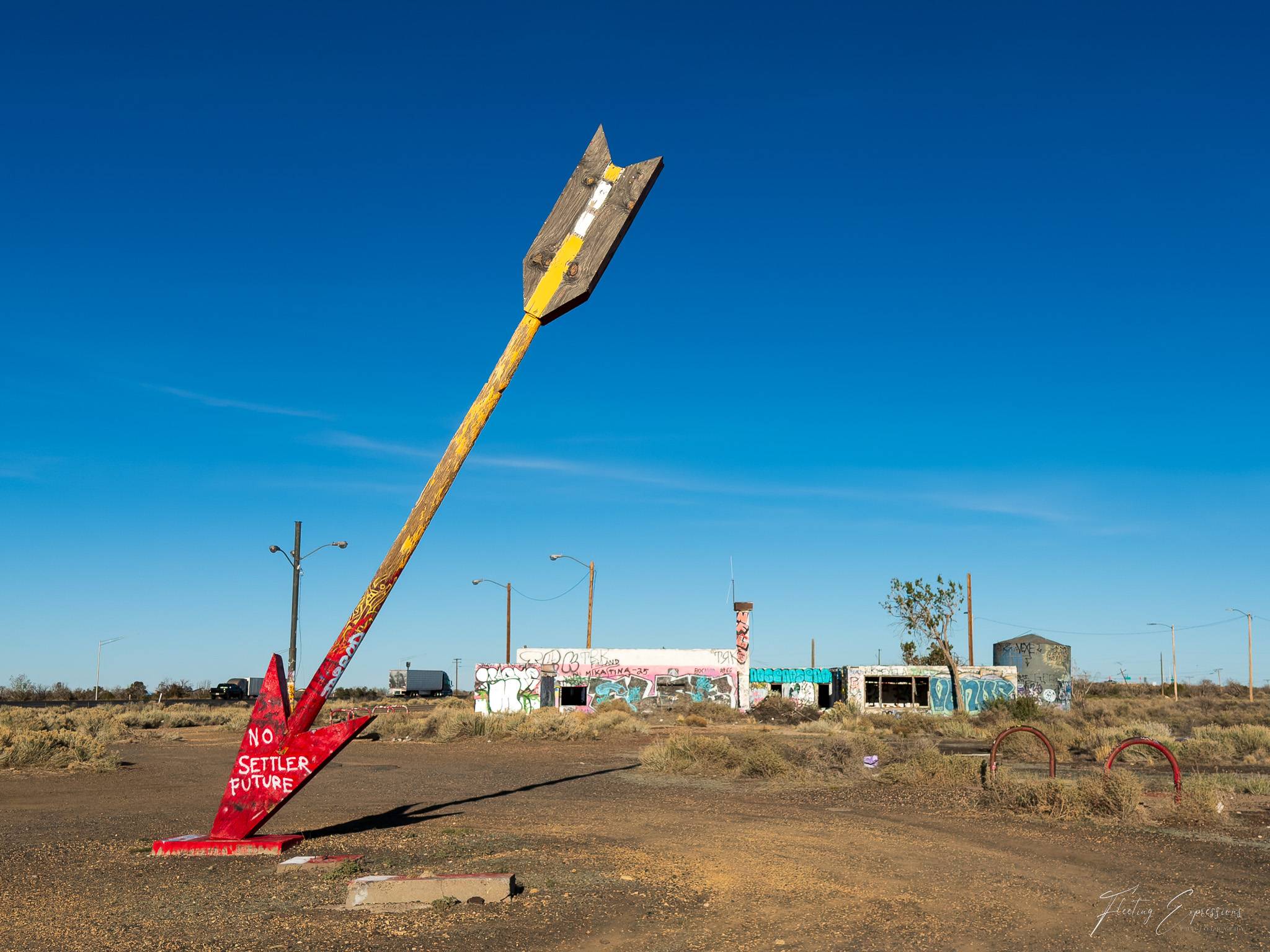 Large arrow sculpture standing in an open desert lot with graffiti-covered buildings and a bright blue sky in the background.