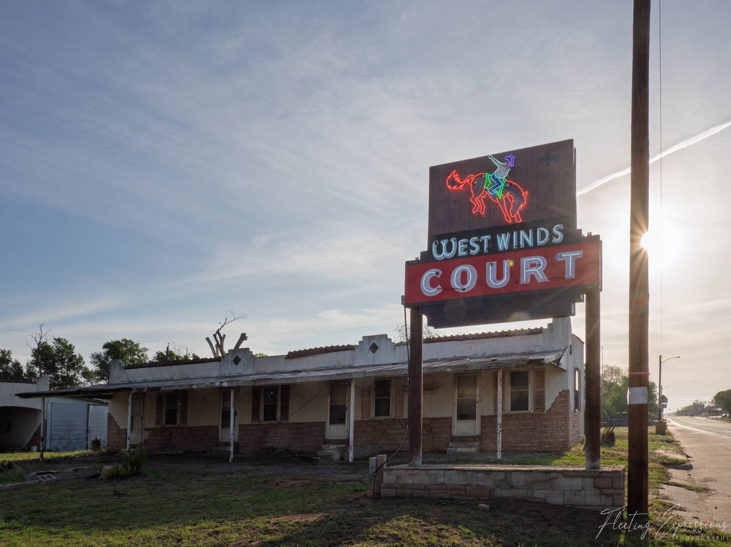 An abandoned single-story brick motel stretches along a quiet Oklahoma highway. A vintage neon sign reading "West Winds Court" with a bucking bronco illustration stands in front of the deteriorating building. The sun flares behind a utility pole in the upper right corner.