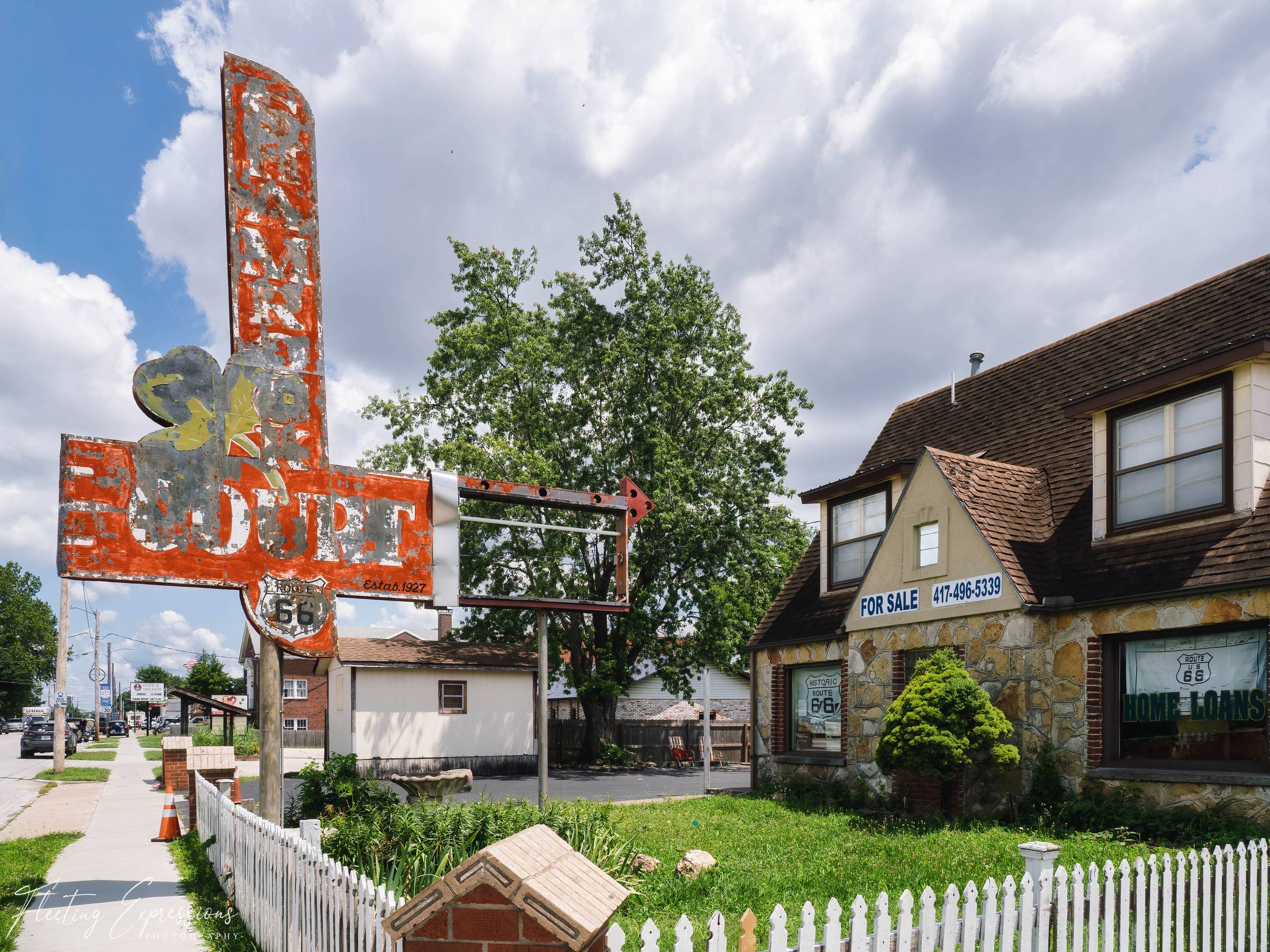 A faded orange Shamrock Court sign marked "Estab. 1927" and topped with a Route 66 shield stands beside a stone cottage with a "For Sale 417-496-5339" sign in the window under a partly cloudy sky.