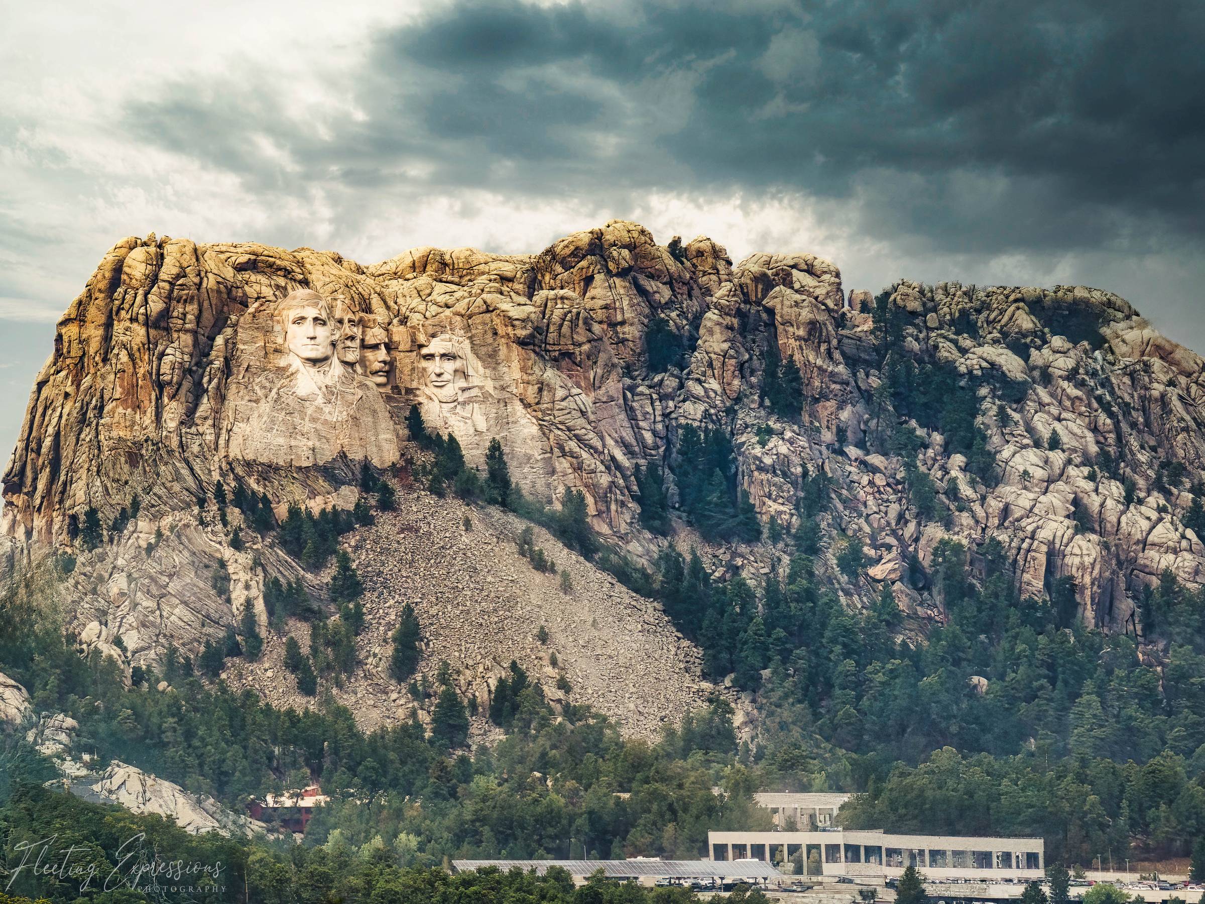 Mount Rushmore's presidential faces lit by golden light against dark, stormy clouds, with pine forest and rocky cliffs surrounding the carving. 