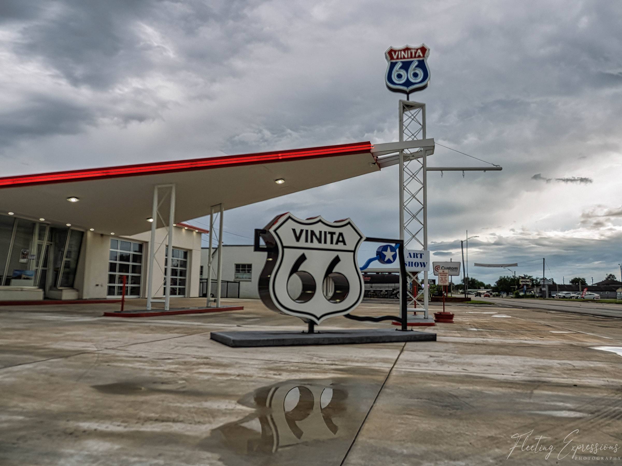 A chrome Route 66 shield reading "Vinita 66" sits on wet pavement beneath a tall neon sign and storm clouds.
