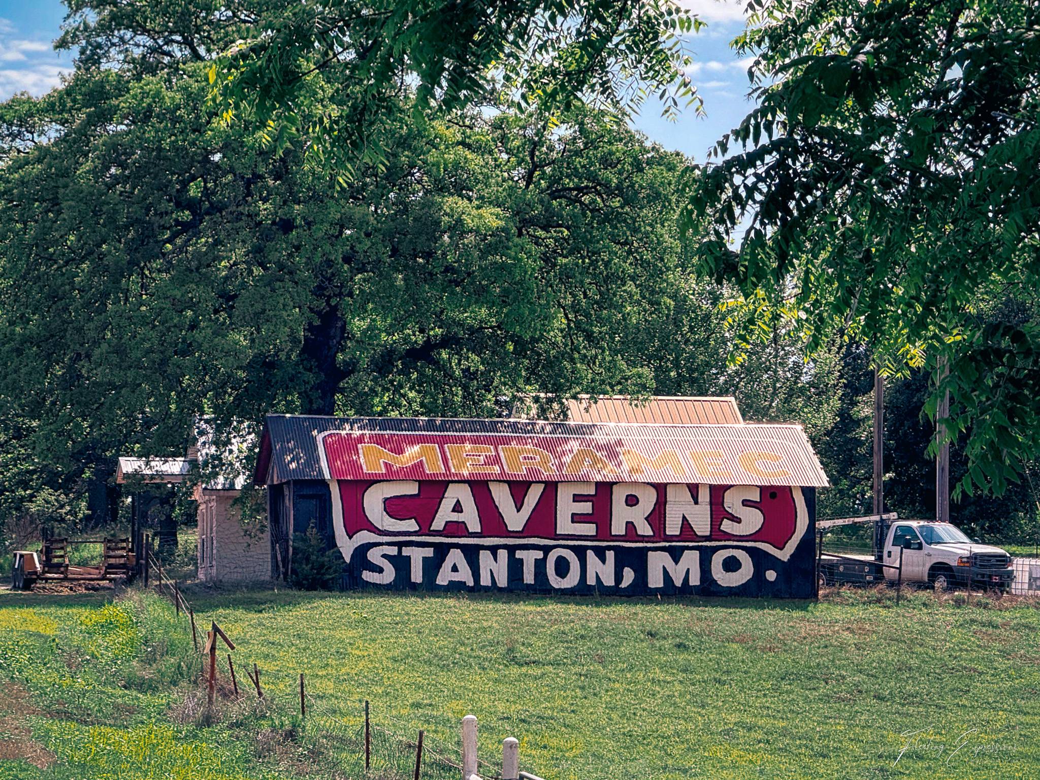 A weathered barn outside Chandler, Oklahoma, with a large faded Meramec Caverns advertisement painted across its side, partially visible beneath a rusty corrugated metal roof and surrounded by green trees.