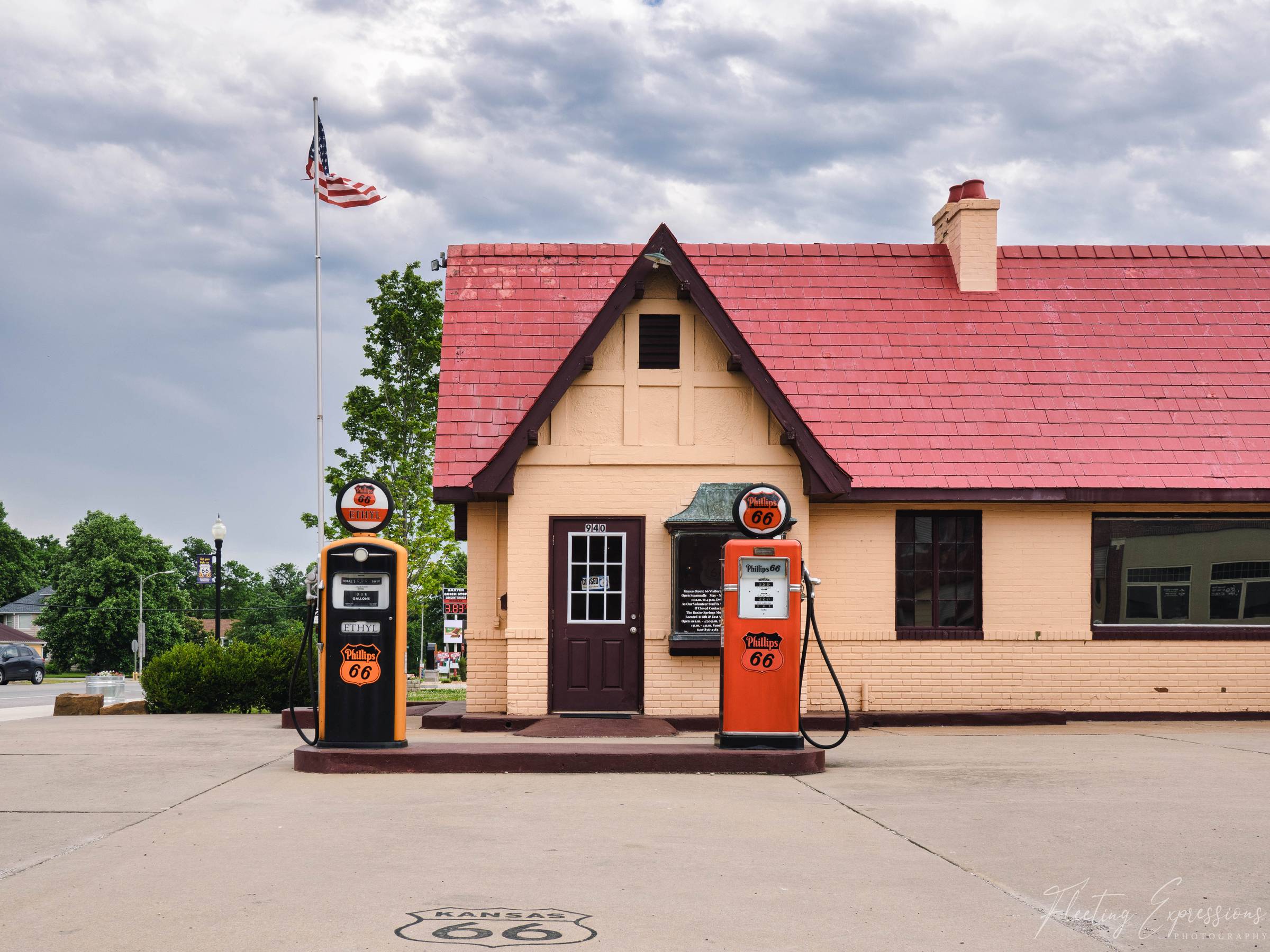 Two restored Phillips 66 pumps stand outside a cottage-style station with a red roof and a Kansas Route 66 shield painted on the pavement.