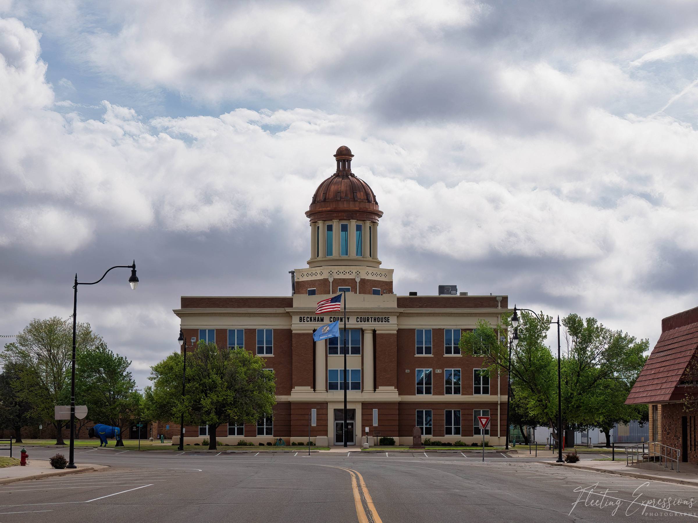 The three-story brick Beckham County Courthouse in Sayre, Oklahoma, topped with a copper dome and flying American and Oklahoma flags, beneath a dramatic cloudy sky over empty streets.
