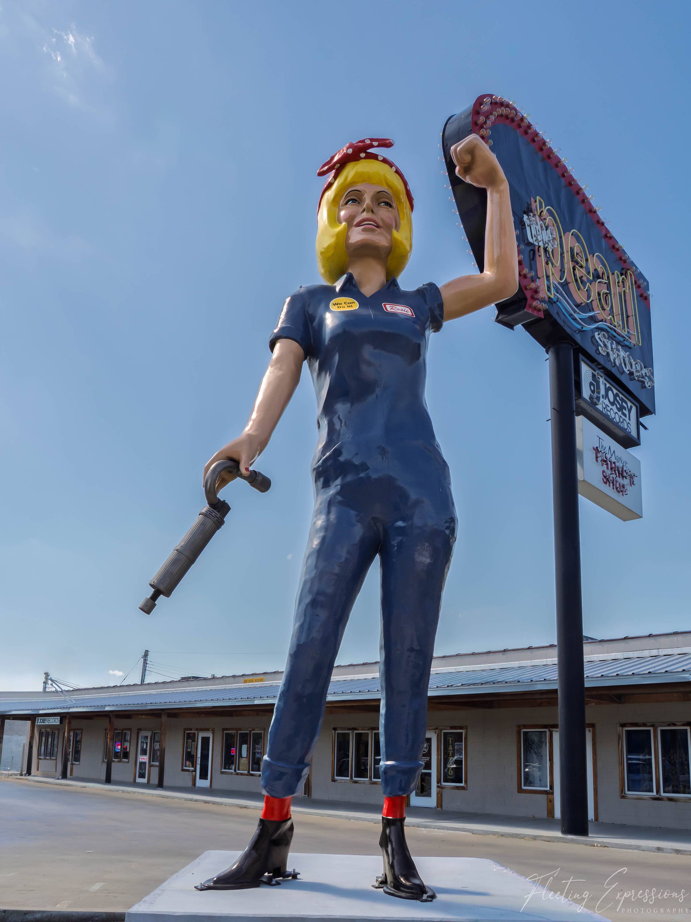 A tall statue of a woman in blue coveralls and a red polka-dot bandana raises her fist beside a neon “Dream” sign. 