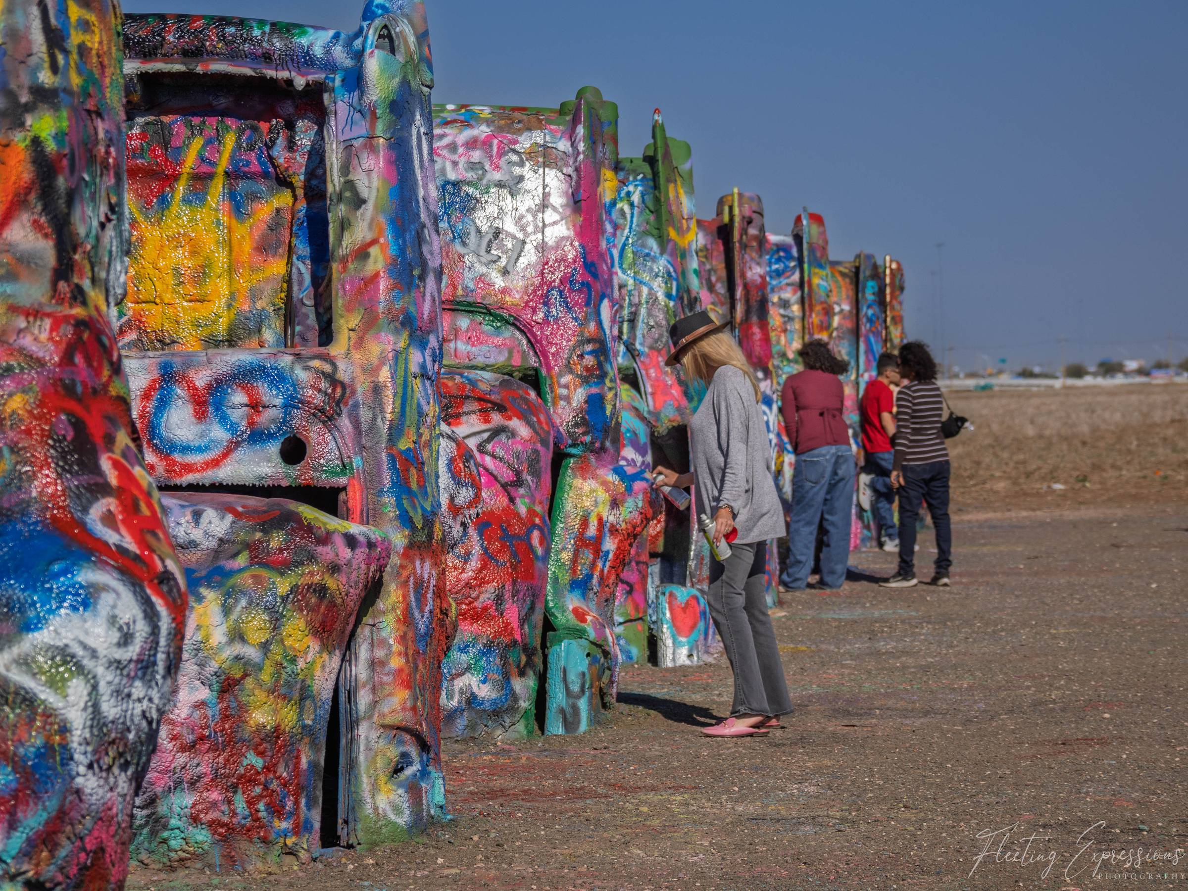 A row of graffiti-covered Cadillacs buried nose-down in a flat Texas field, with visitors adding their own paint in the afternoon sun.