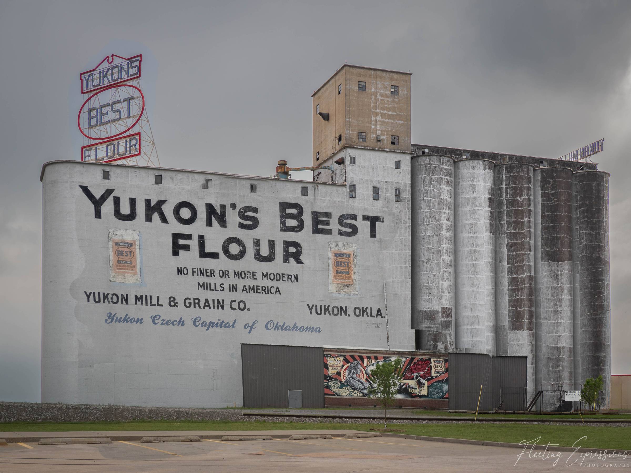 The historic Yukon Mill and Grain Company silos painted white with large black lettering reading Yukon's Best Flour, with a vintage neon sign visible against a stormy sky in Yukon, Oklahoma.